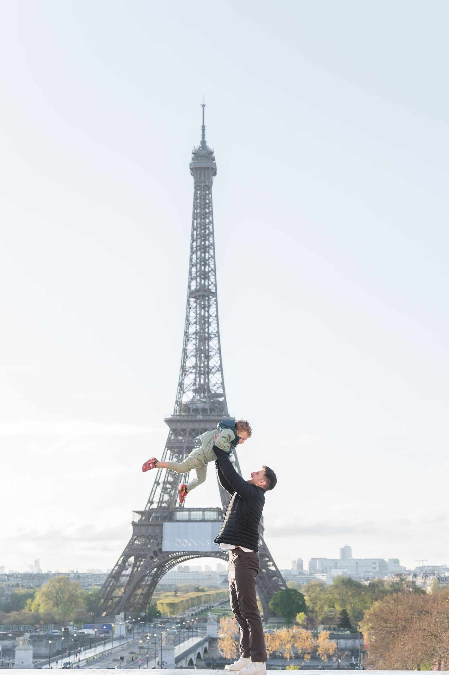 A father joyfully lifts his child in front of the Eiffel Tower in Paris on a clear autumn day.