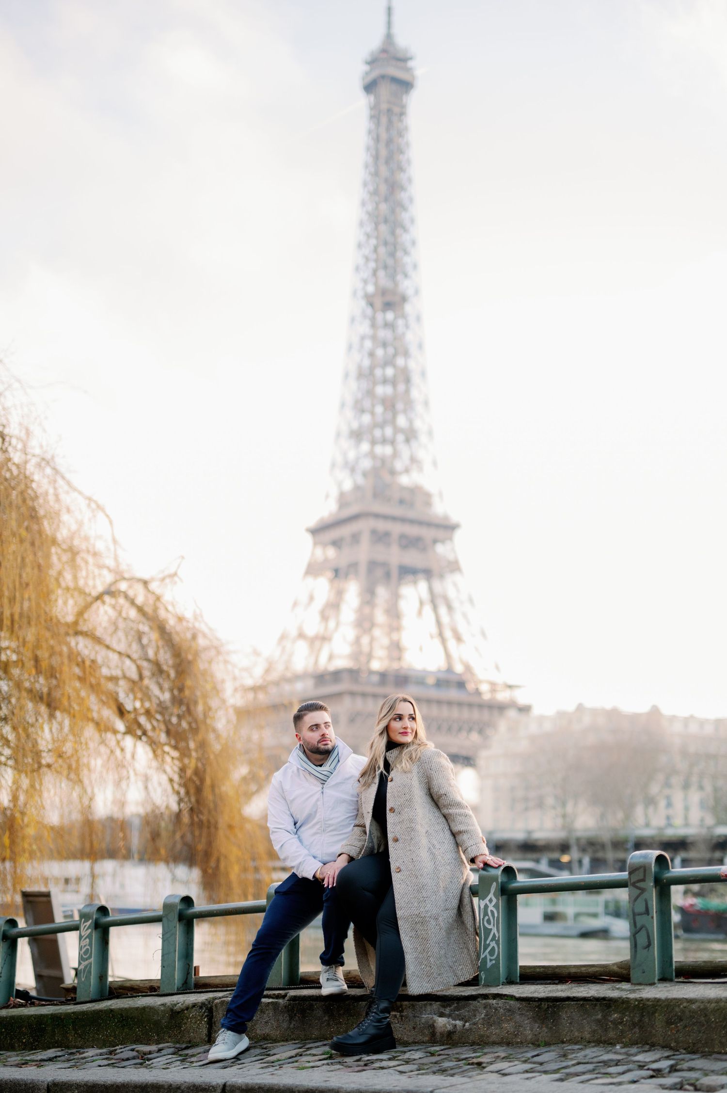 A couple poses together near the Seine River with the iconic Eiffel Tower in Paris during winter.