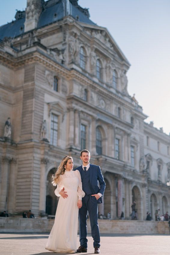 A couple in wedding attire stands together in front of the grand Louvre Museum in Paris on a sunny day.
