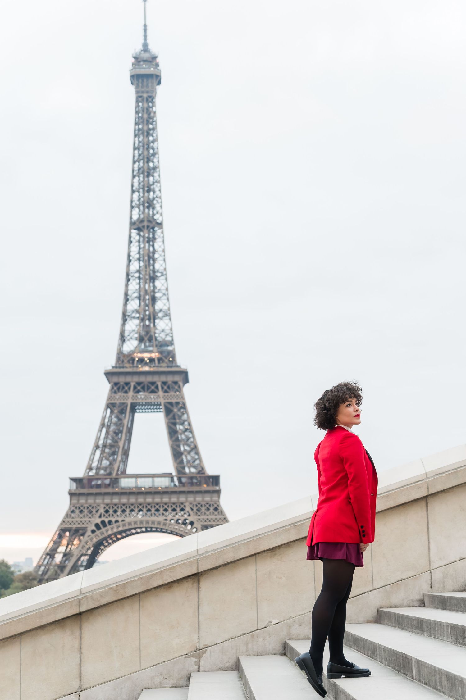 A person in a red coat poses on the steps of Trocadero with the Eiffel Tower visible in the background.