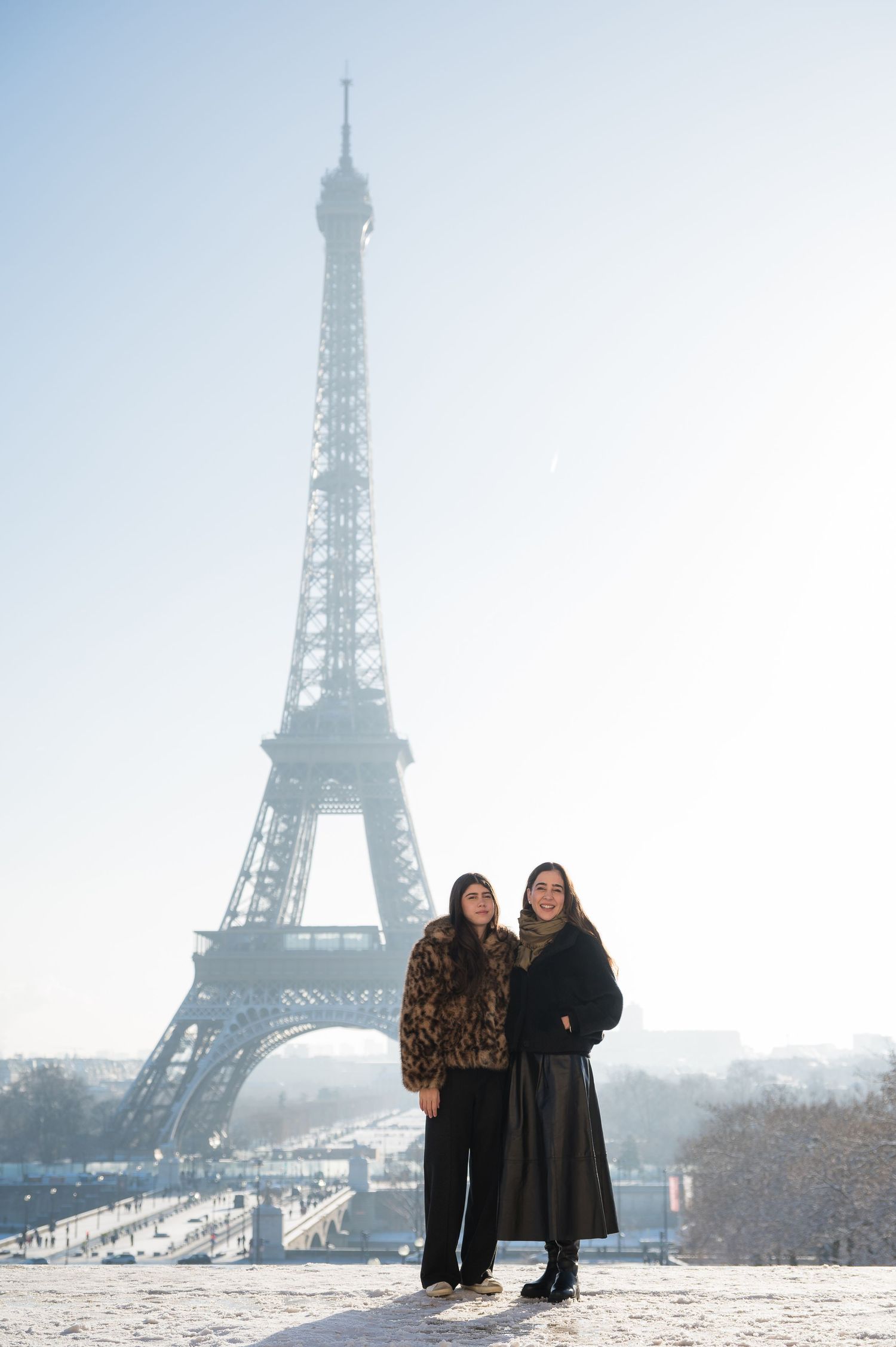 Two people pose together in winter coats against the backdrop of the Eiffel Tower on a snowy winter day in Paris.