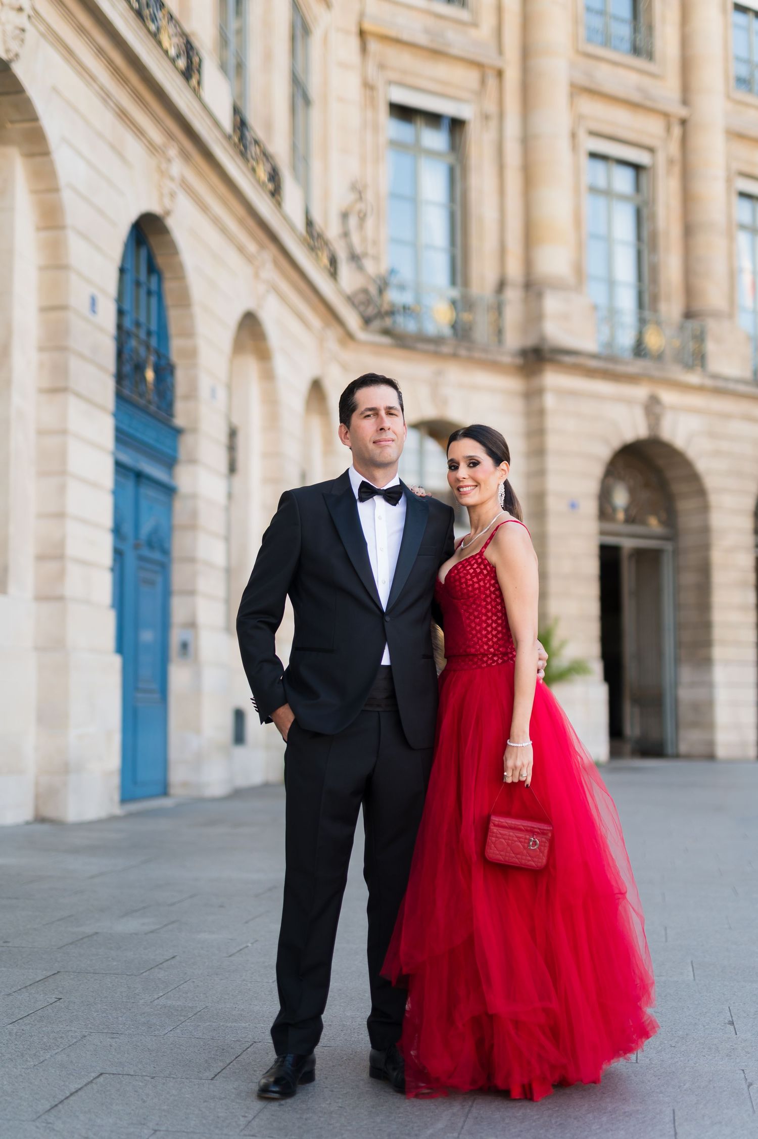 Elegant couple posing in Paris, woman in red tulle gown with beaded bodice, man in black tuxedo with bow tie.