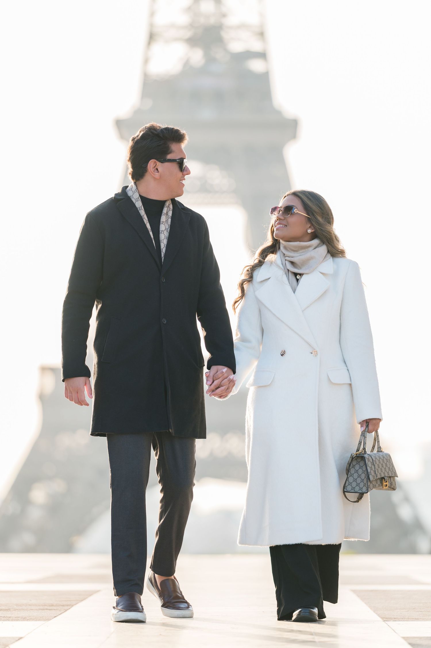 Couple holding hands walking near the Eiffel Tower in Paris, wearing elegant white and black coats on a sunny day.
