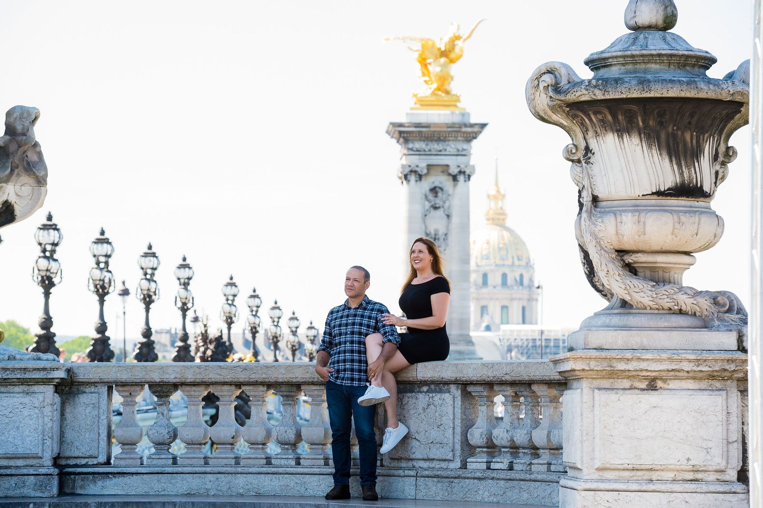 A couple stands on the iconic Alexandre III bridge in Paris with ornate lamp posts and the golden Invalides dome in background.