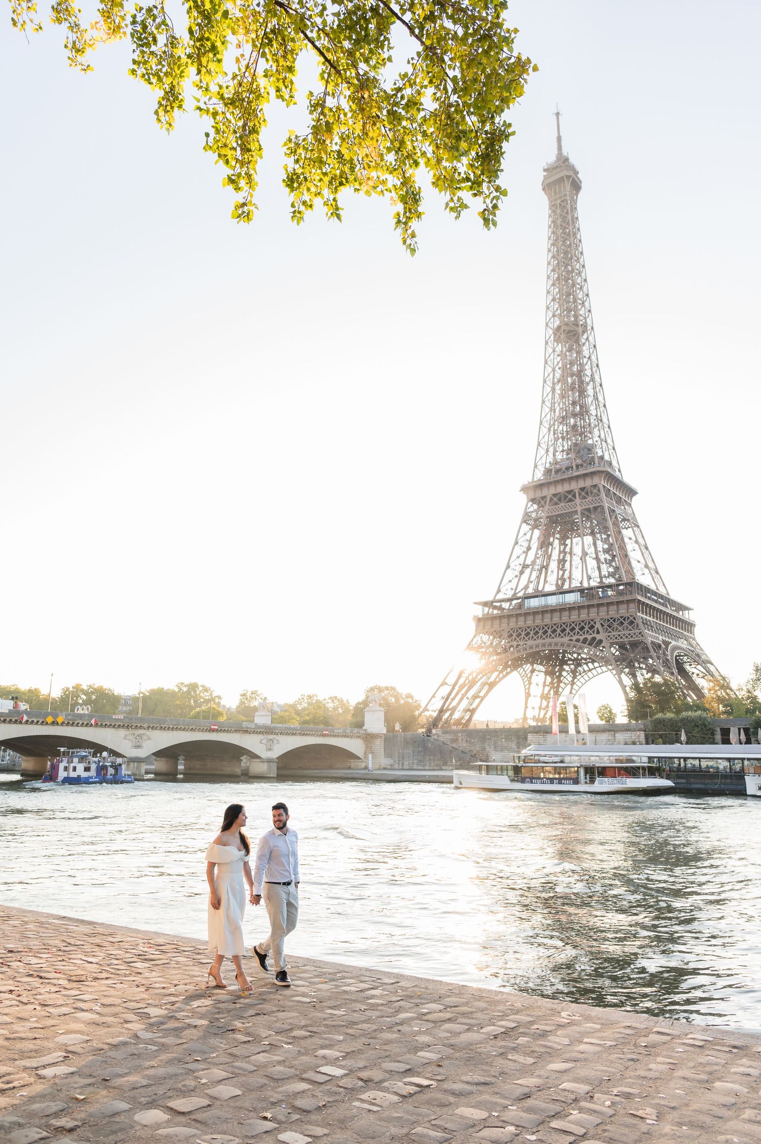 A couple strolls along the Seine River with the iconic Eiffel Tower in the background during sunset.