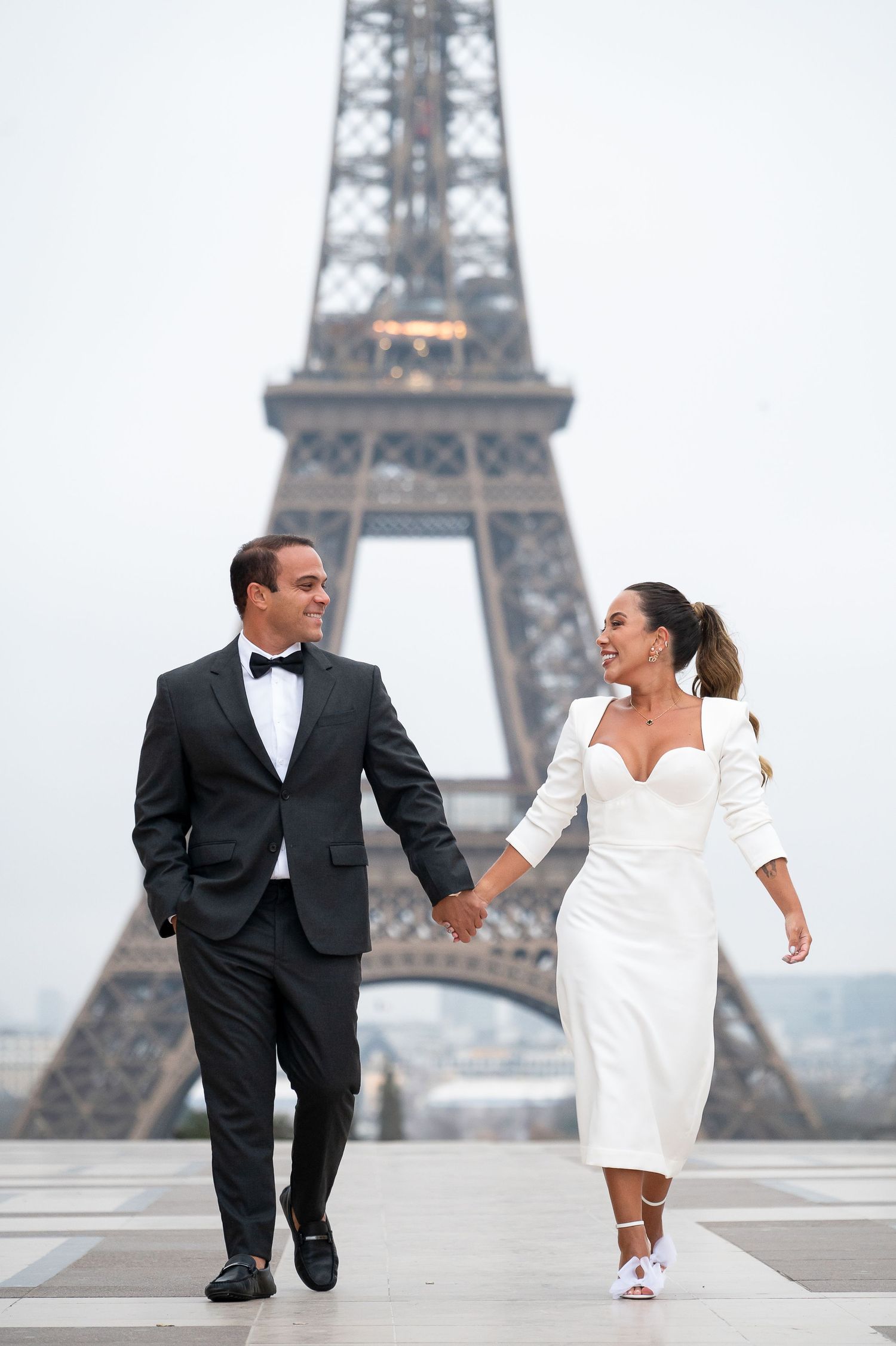 Elegant couple in formal attire walking hand in hand by the Eiffel Tower.