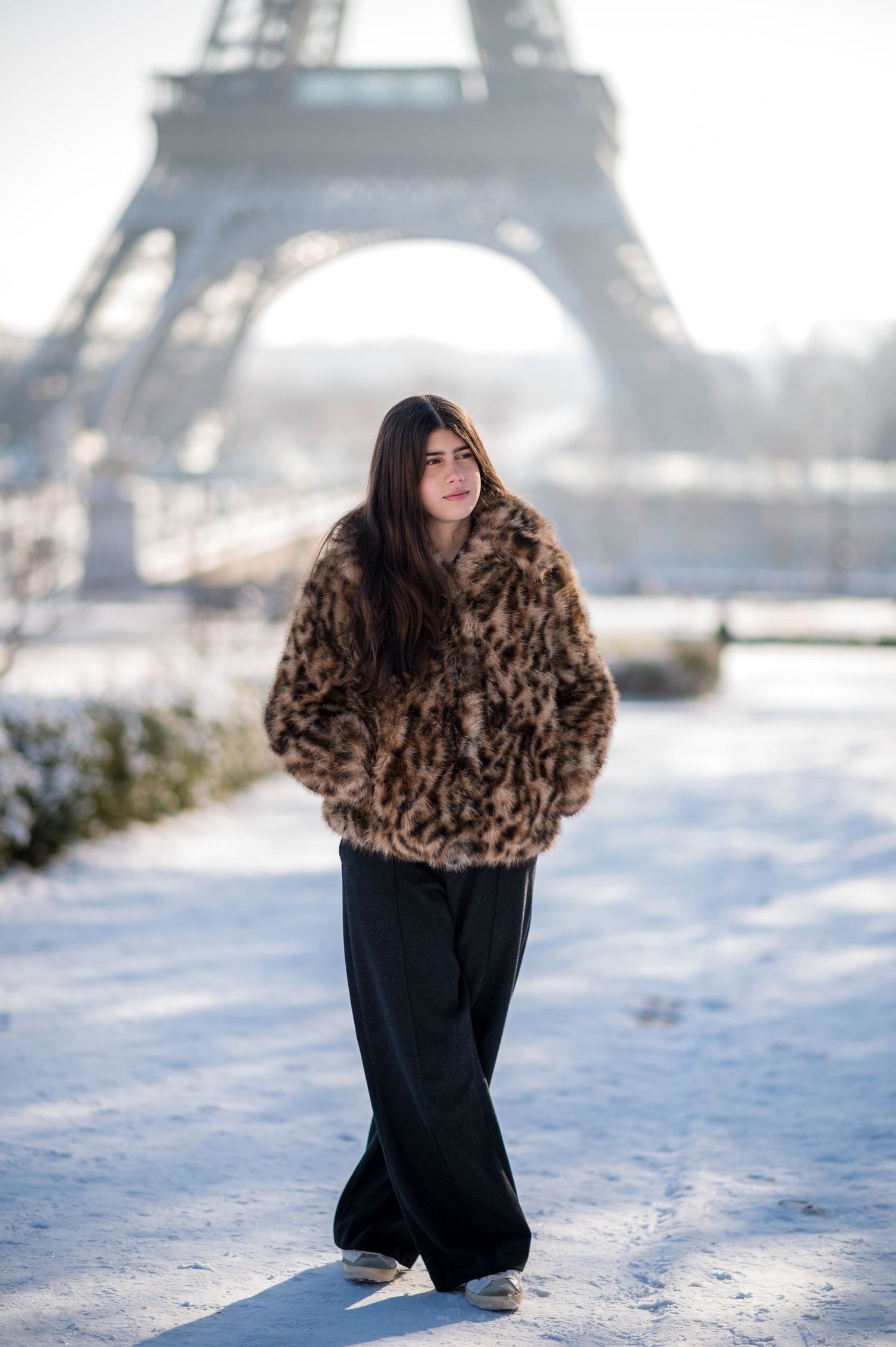 A stylish winter outfit with brown fur coat and wide-leg pants against snowy Paris with the Eiffel Tower in background.