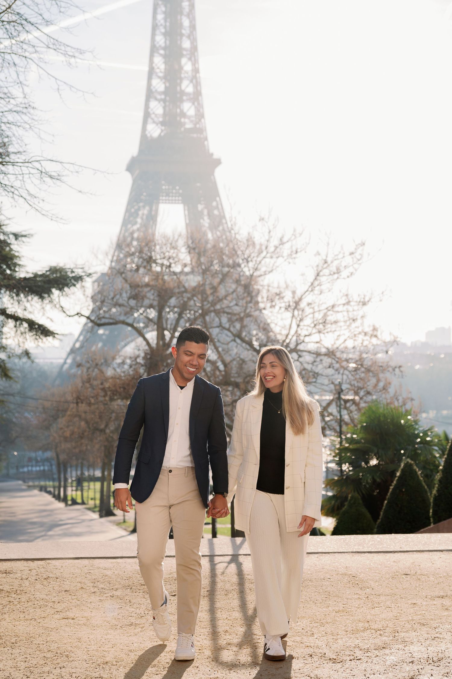 A stylish couple in matching white outfits poses on a path with the Eiffel Tower backdrop in Paris.