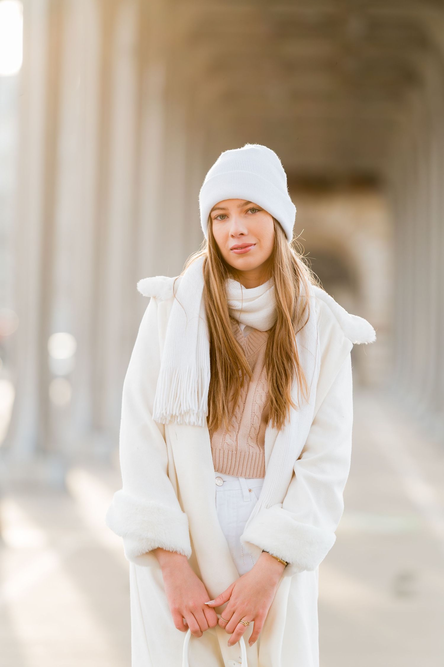 Cozy winter fashion ensemble with white knit beanie and cream coat in architectural setting.
