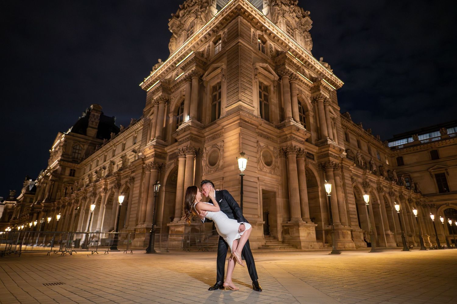 A romantic couple shares a passionate embrace outside the illuminated Louvre Museum in Paris at night.