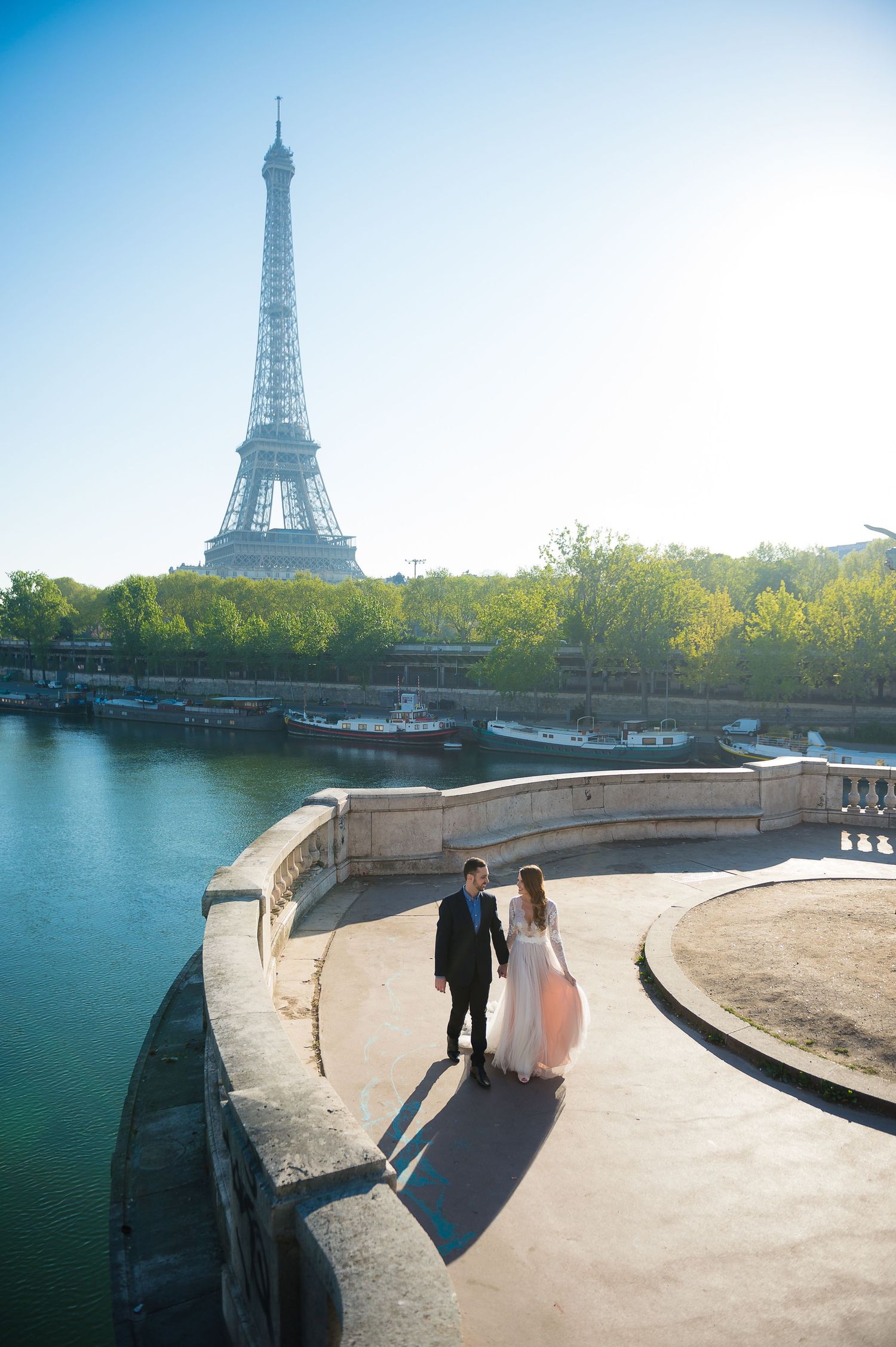 Wedding couple walking along Seine riverbank with iconic Eiffel Tower rising behind them in Paris.