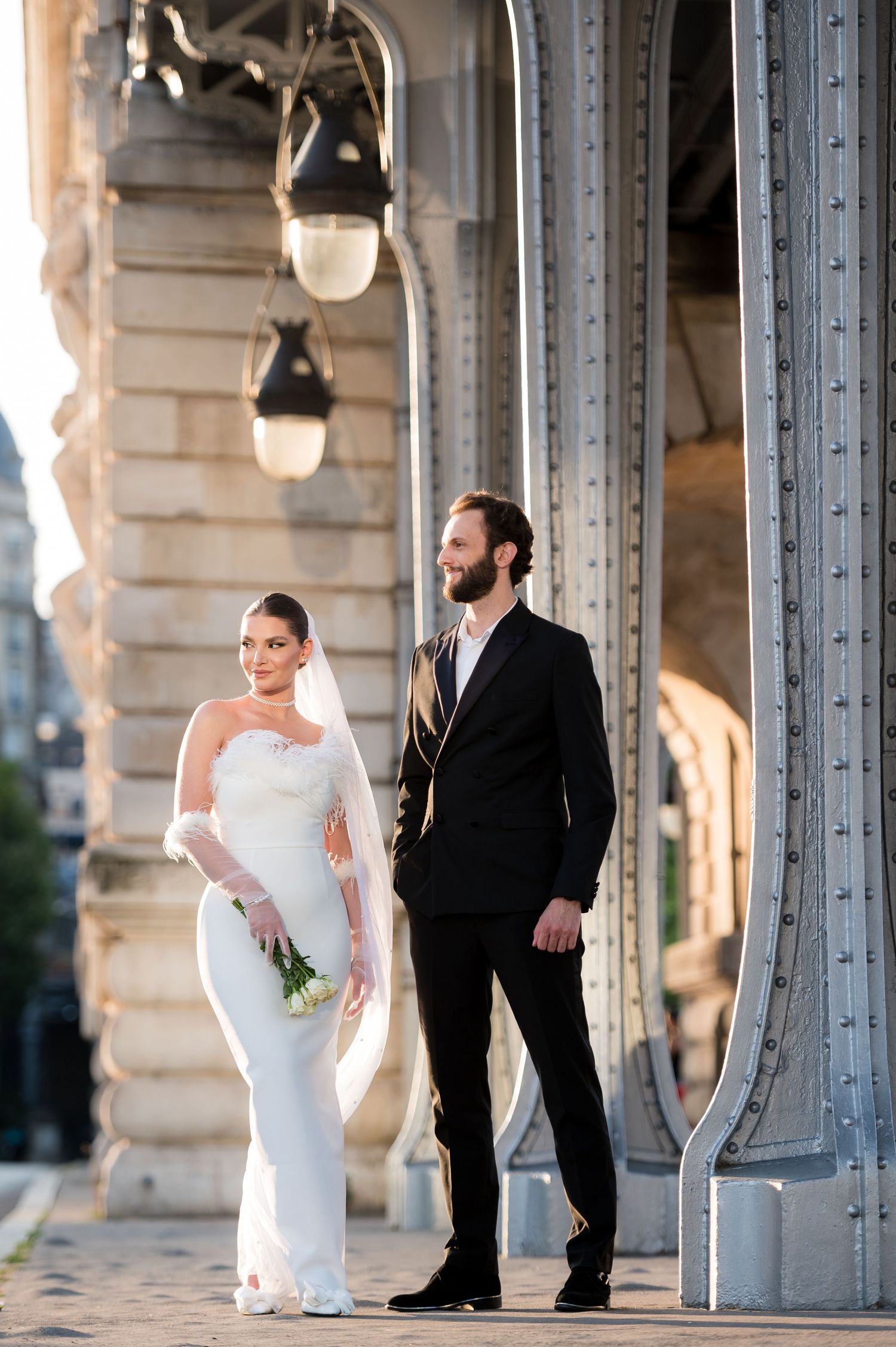 A newlywed couple poses elegantly on a Parisian bridge with ornate architecture and vintage lampposts in the background.
