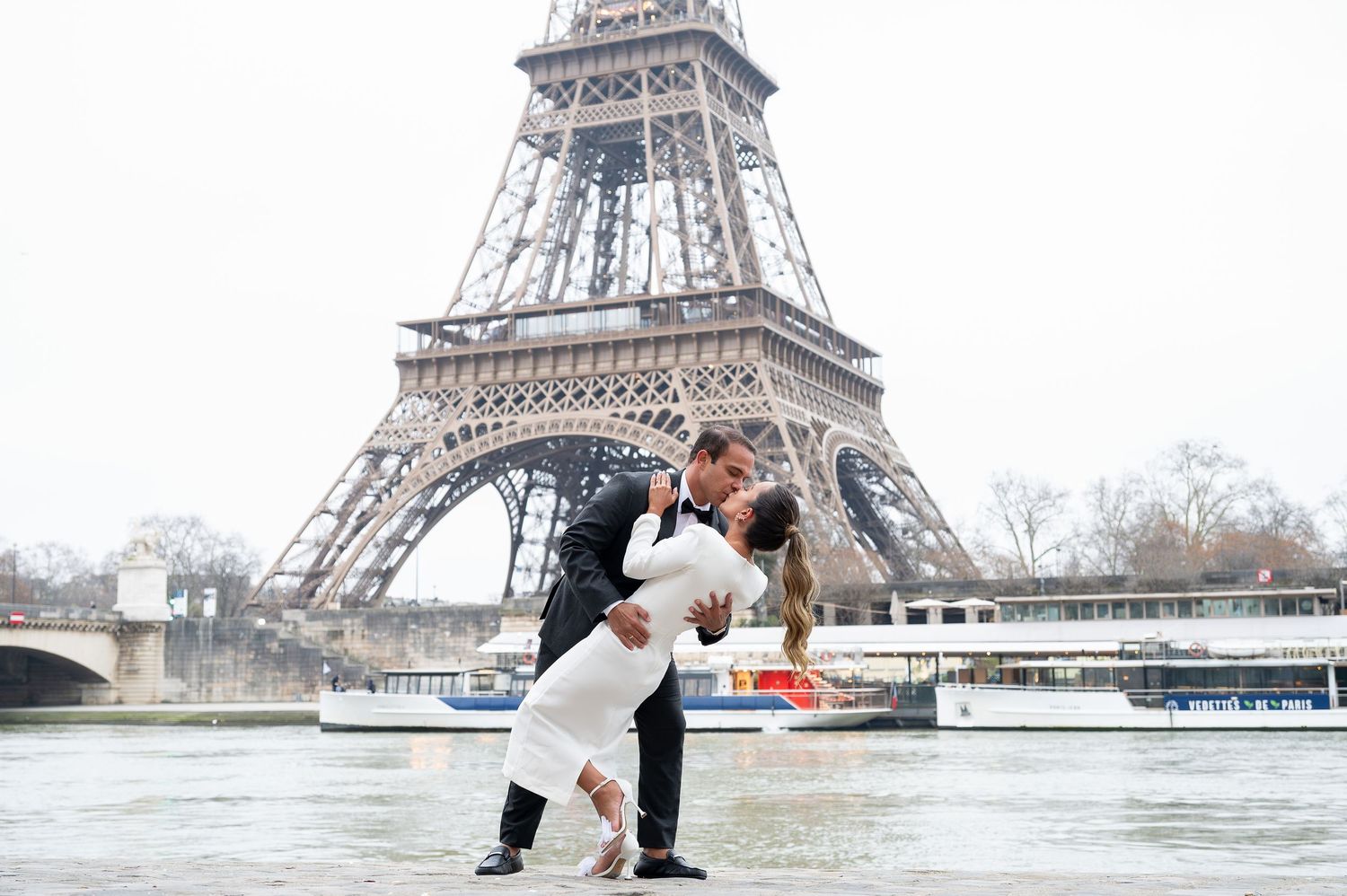 A romantic couple embraces near the iconic Eiffel Tower in Paris on a cloudy winter day.