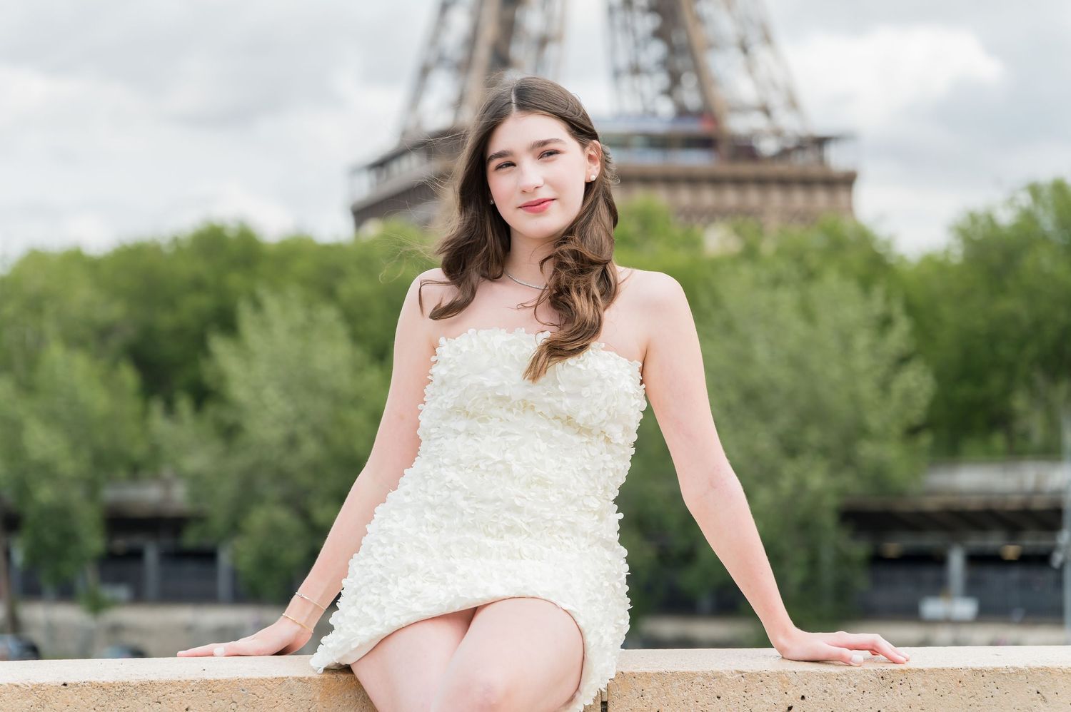 Bride in white wedding dress poses gracefully along Seine River with iconic Eiffel Tower visible in Paris skyline.