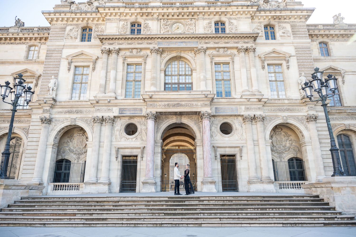 A wide view of a couple on the steps of a classical French building with ornate stone architecture.