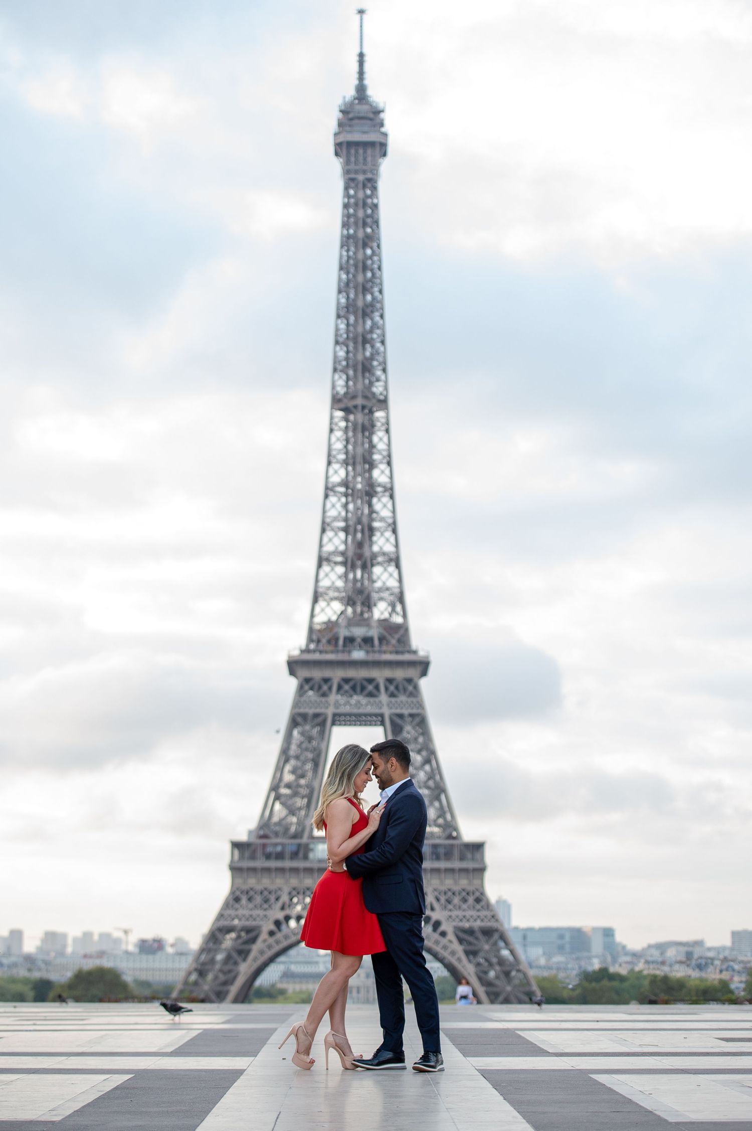 Couple embracing romantically in front of the Eiffel Tower in Paris, woman in red dress, man in dark suit.