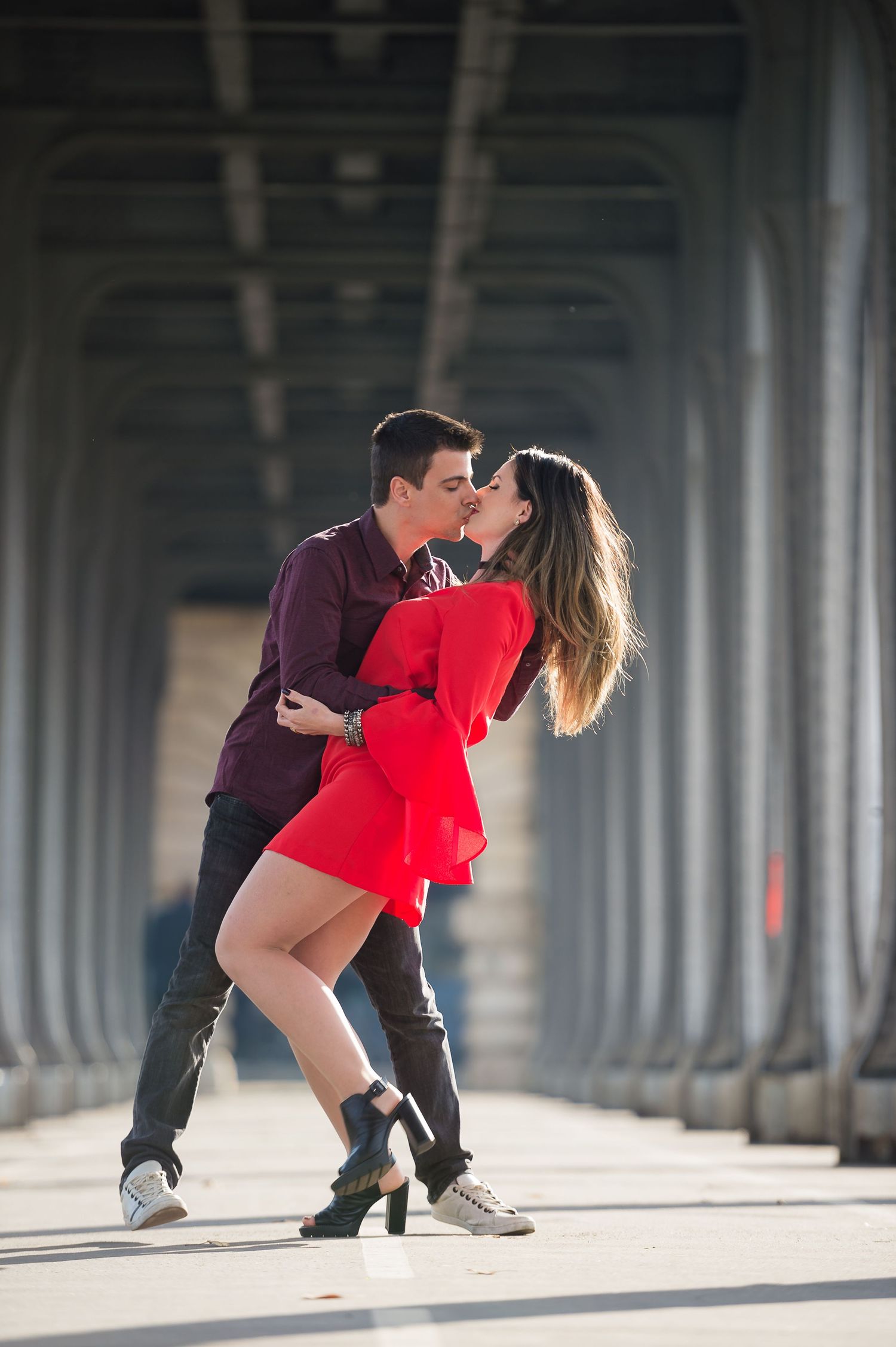 A romantic couple shares an intimate dance pose beneath an industrial bridge while the woman wears a striking red dress.