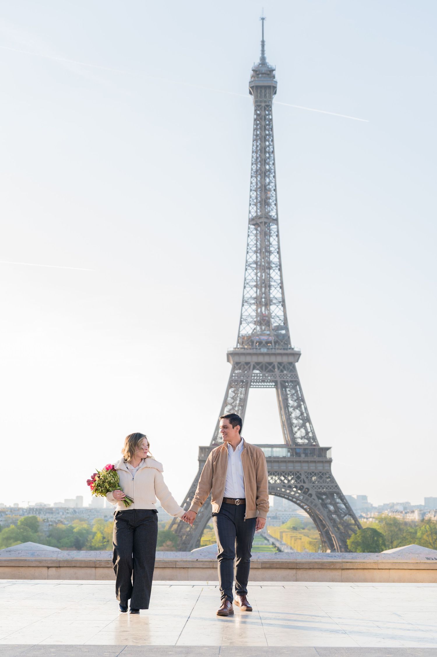 Couple holding hands and walking with flowers near Eiffel Tower in soft morning light.
