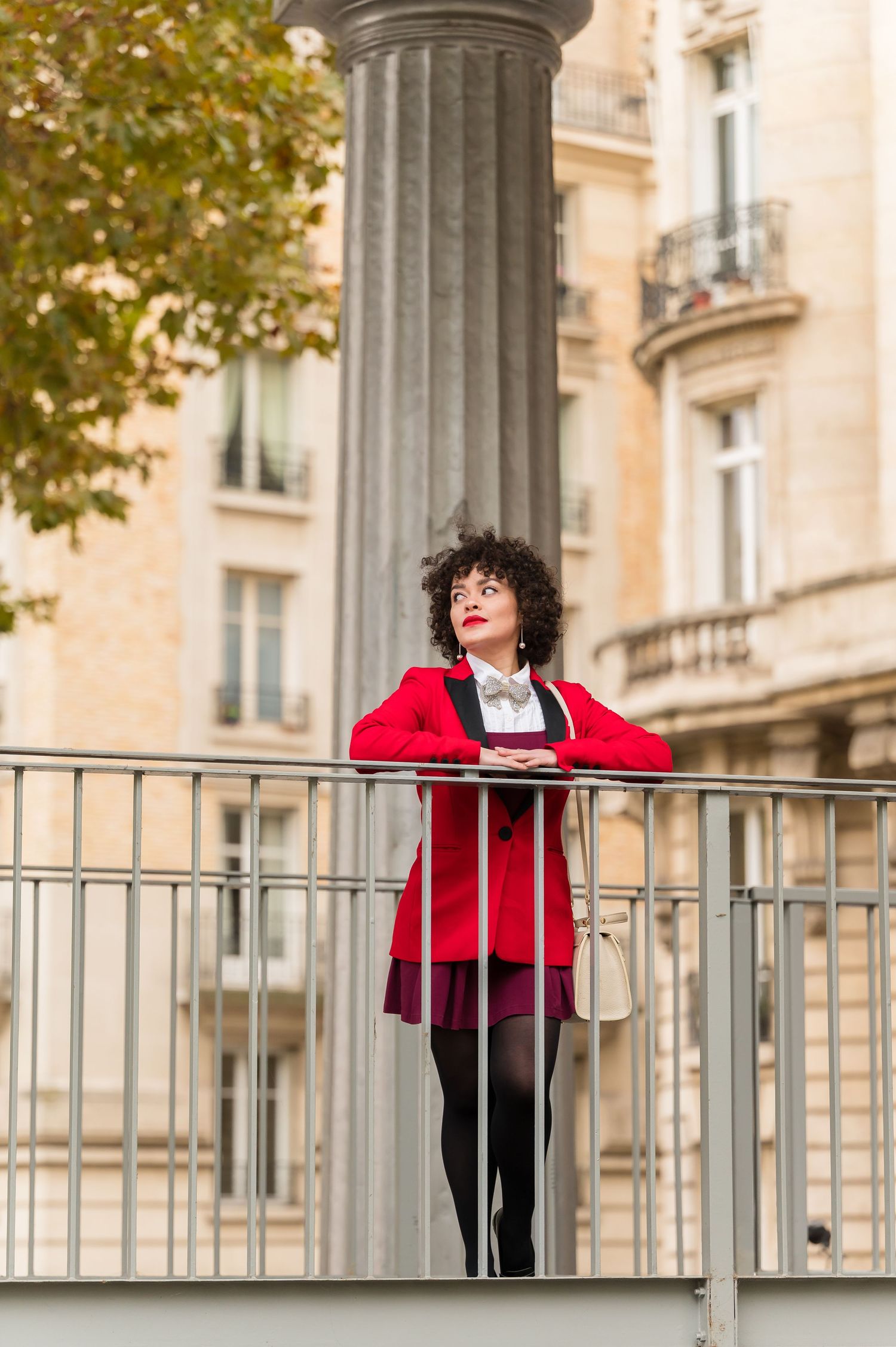 A person in a red cardigan and black outfit poses playfully on a balcony railing with classic architecture in the background.
