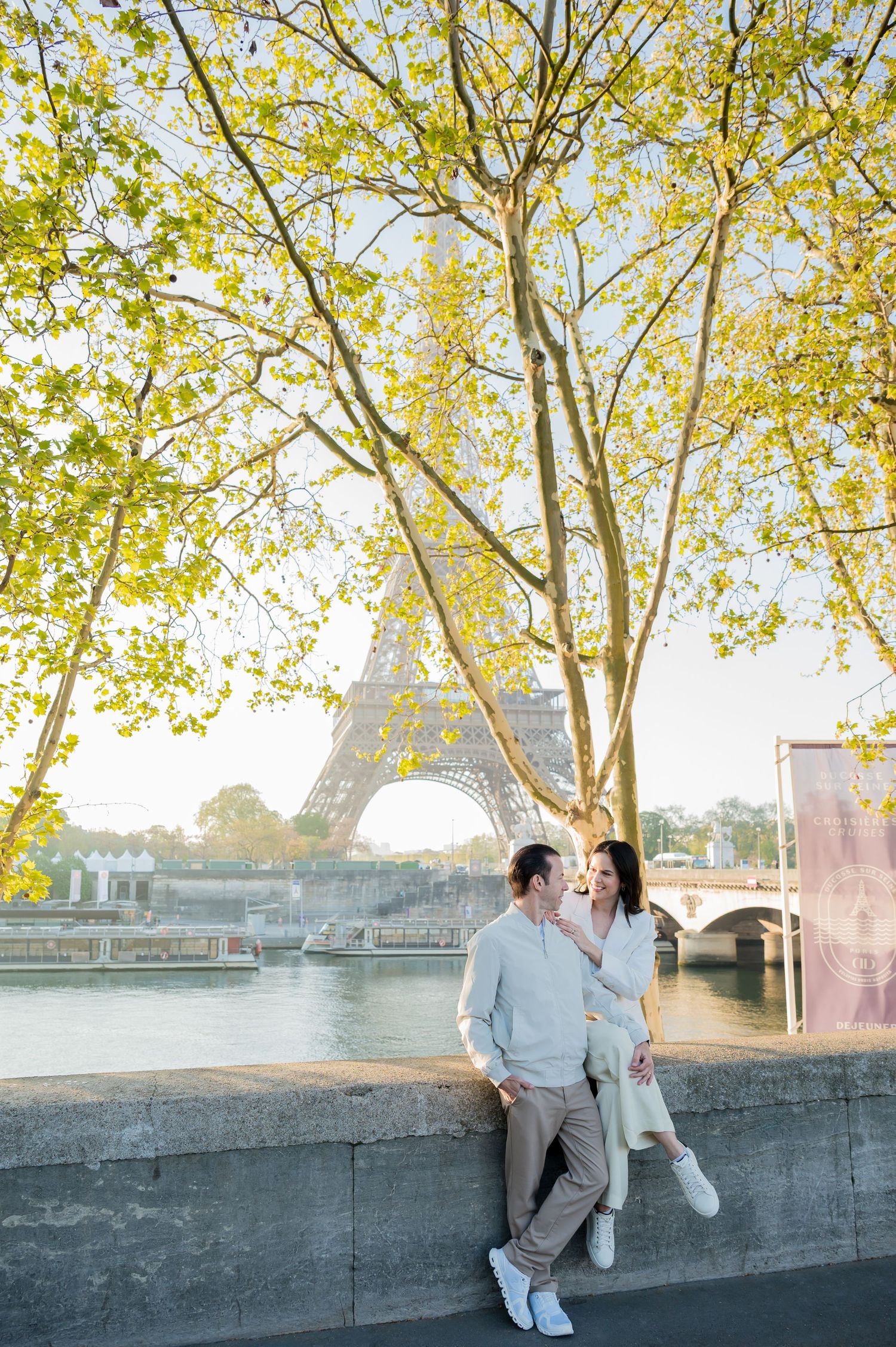 A couple shares a romantic moment along a riverside wall with autumn trees and water in the background.