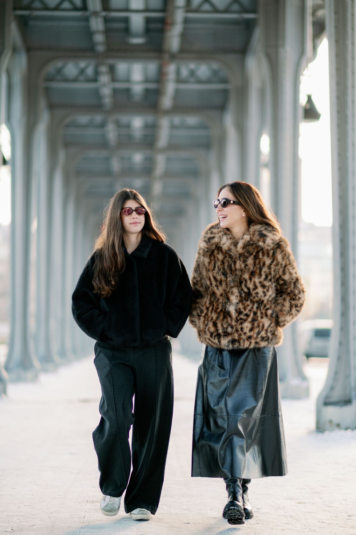 Two people in winter coats walk and chat together under an ornate covered walkway with arched architecture.