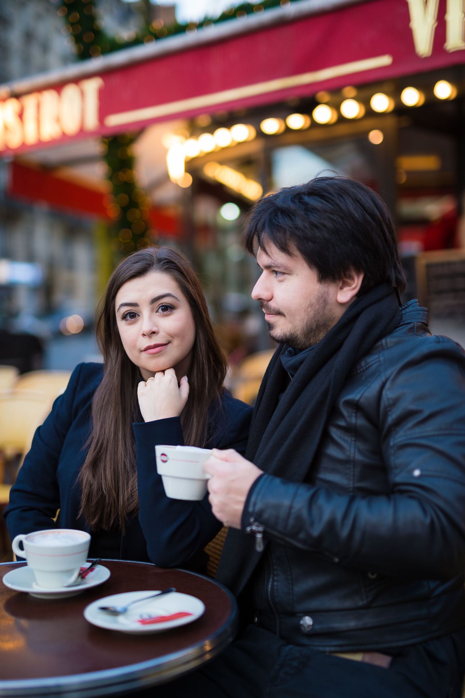 A couple enjoys coffee at an outdoor Parisian cafe with glowing lights in the background.
