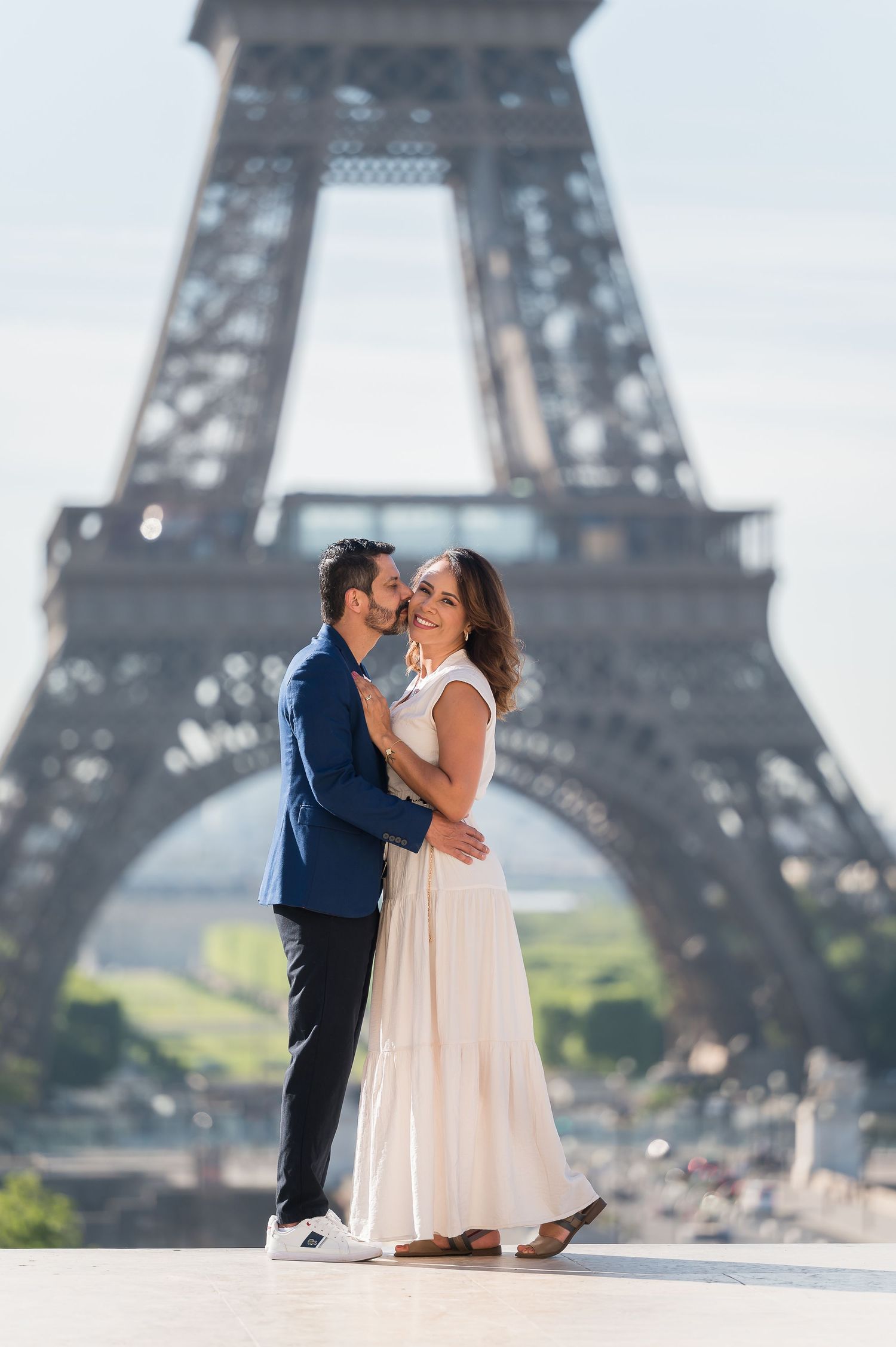 Couple embracing romantically in front of the Eiffel Tower in Paris, woman in white dress, man in blue blazer.