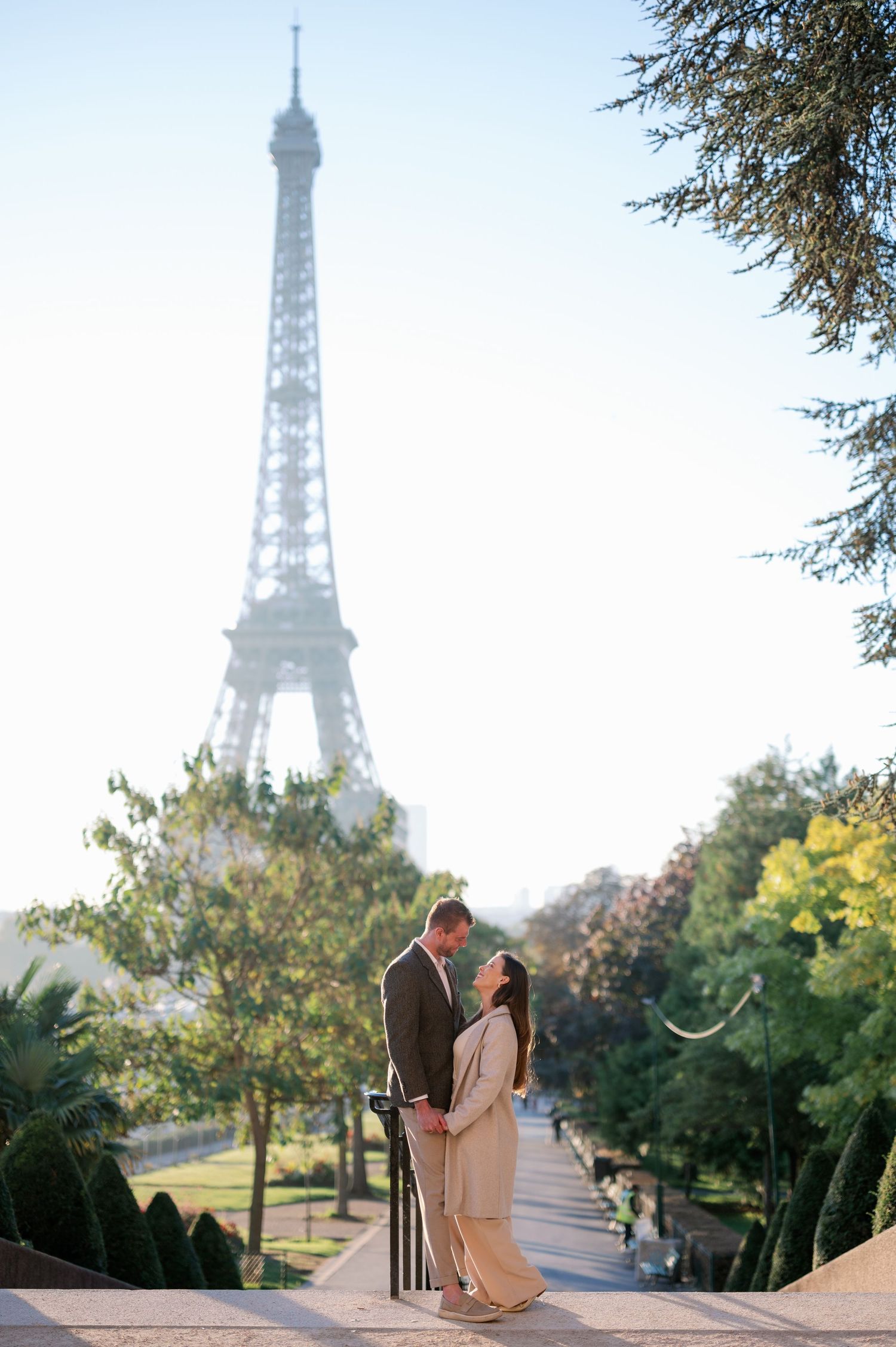 Couple embracing romantically in front of the Eiffel Tower in Paris, surrounded by lush green trees on a sunny day.