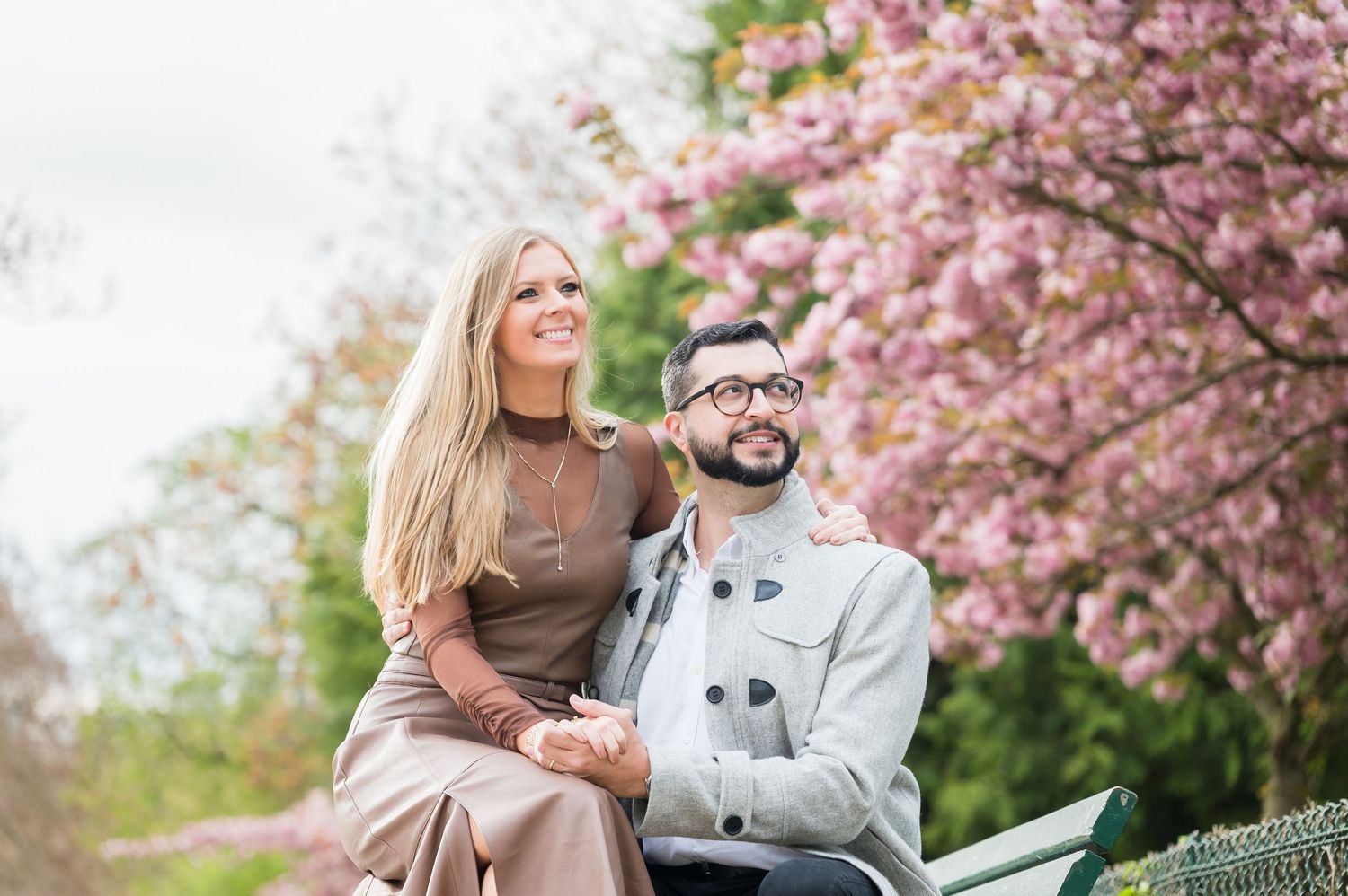 Couple sitting on a bench under pink cherry blossom trees in a park, smiling and holding hands.