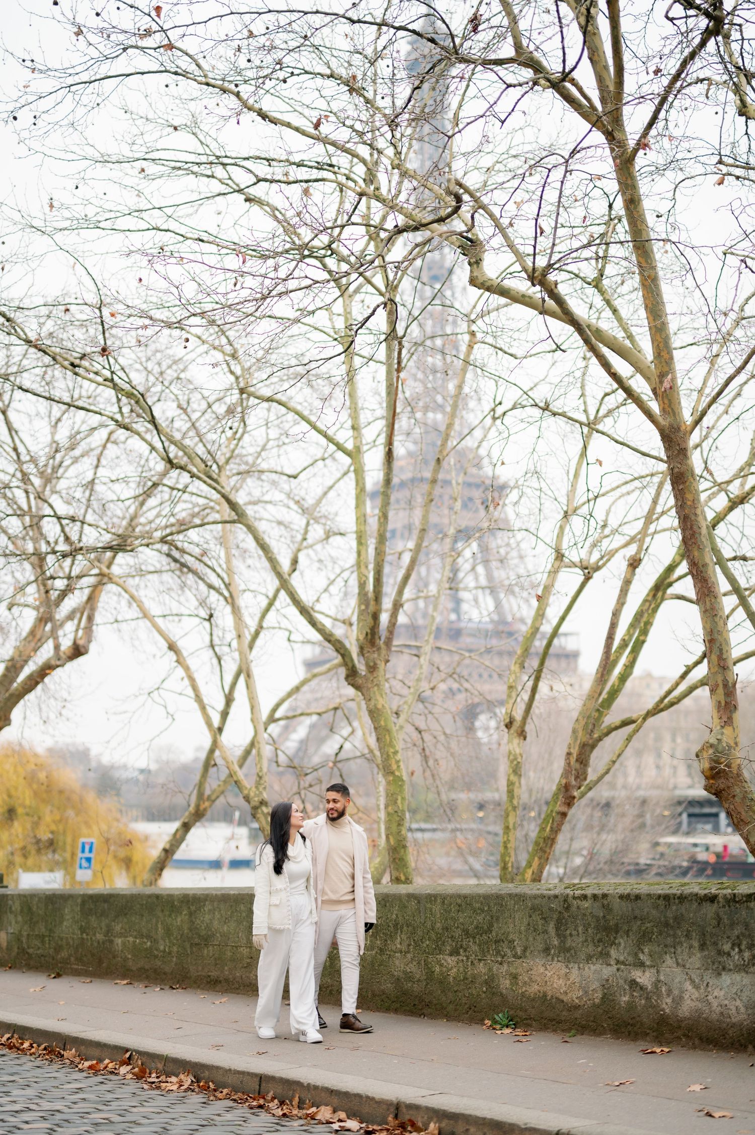 A couple in matching cream outfits stands on a sidewalk near bare trees with the Eiffel Tower visible in the misty background.