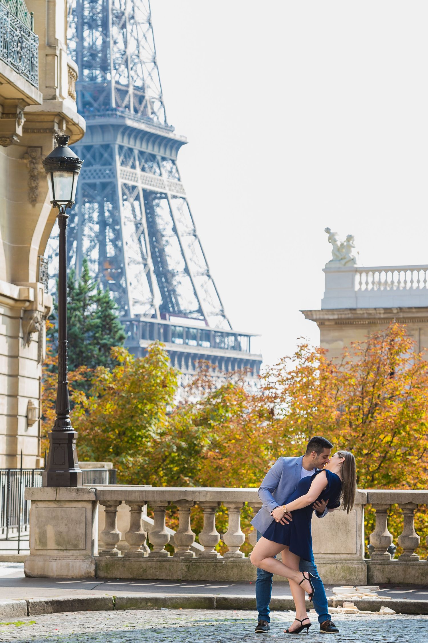 A couple embraces near the Eiffel Tower in Paris during autumn with yellow-leafed trees in the background.