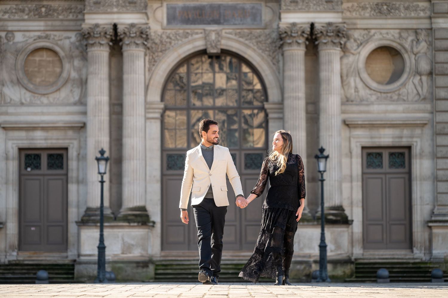 A couple stands in formal attire outside a grand Parisian building with classical architecture.