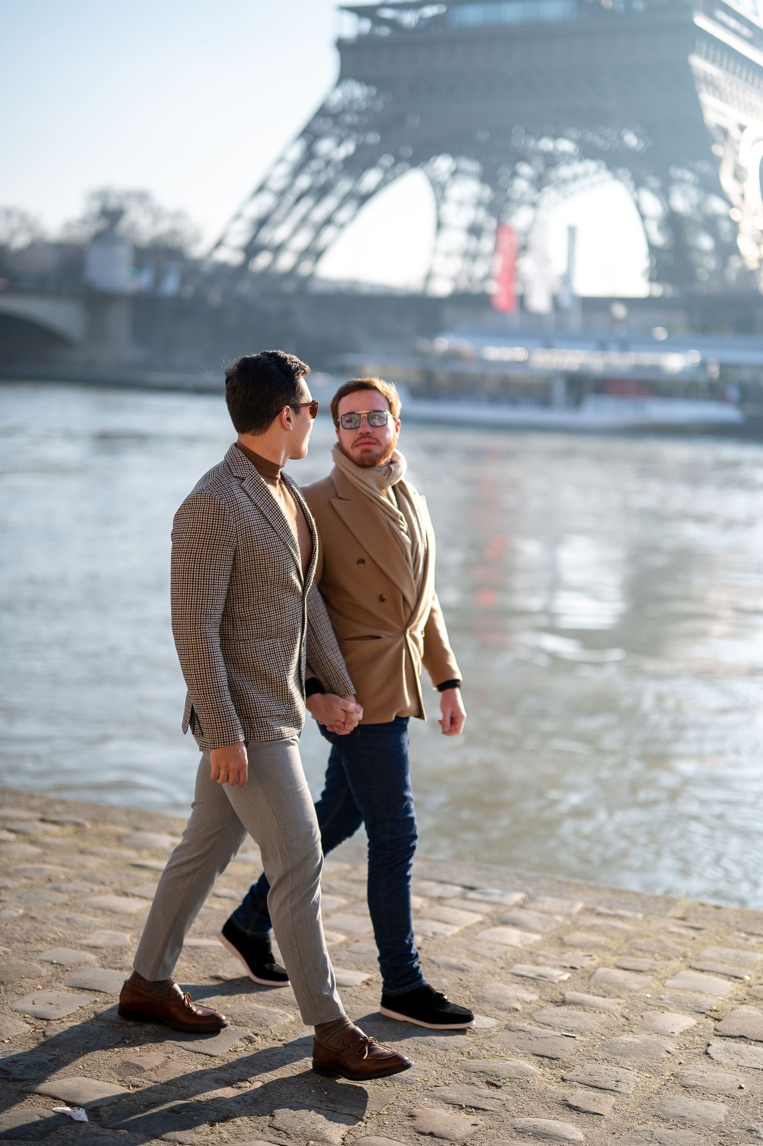 Two stylishly dressed people walking along the Seine River with the Eiffel Tower visible in the background.