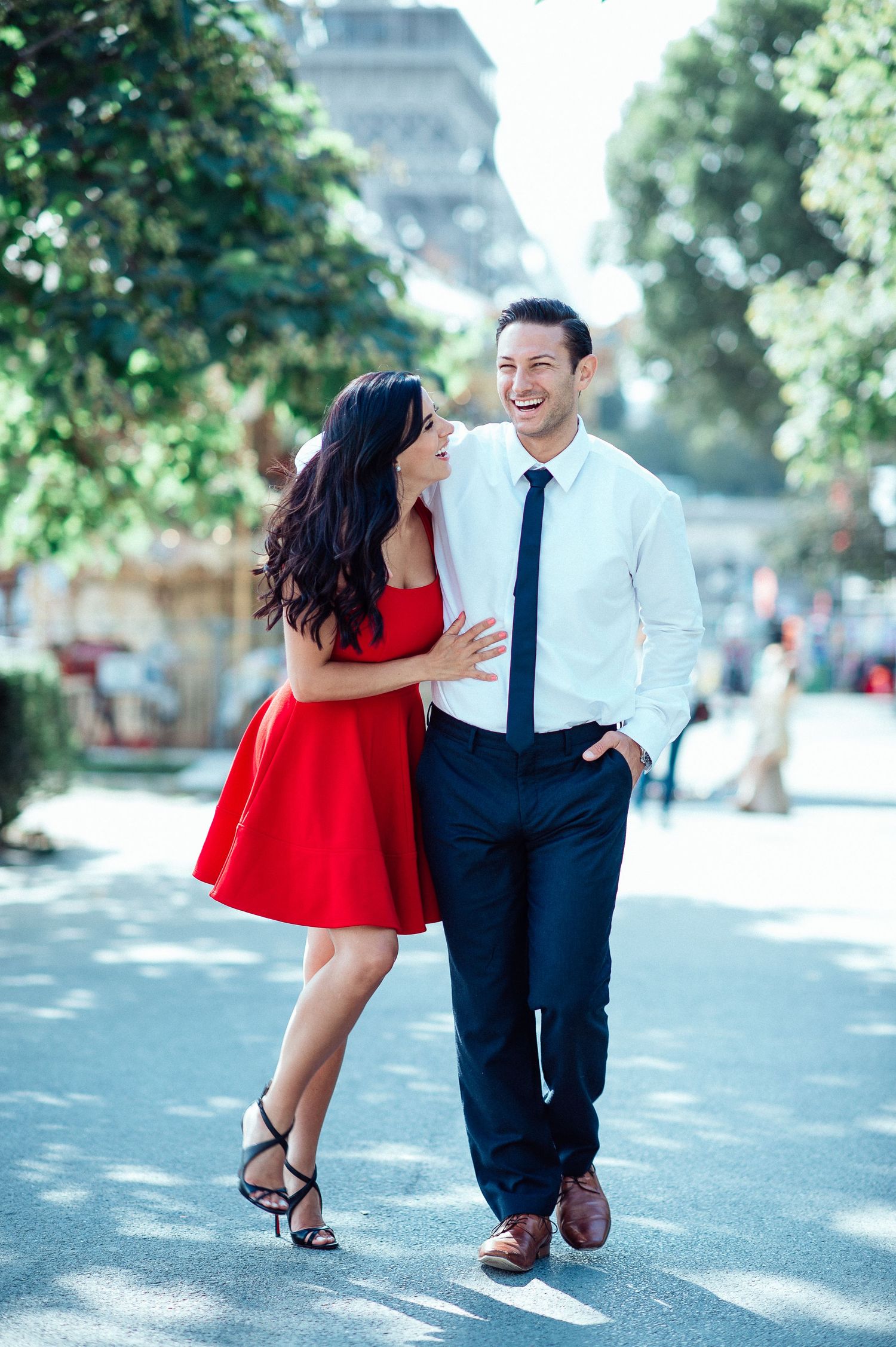 A smiling couple walks together in Paris, woman in red dress, man in white shirt and tie, Eiffel Tower in background.
