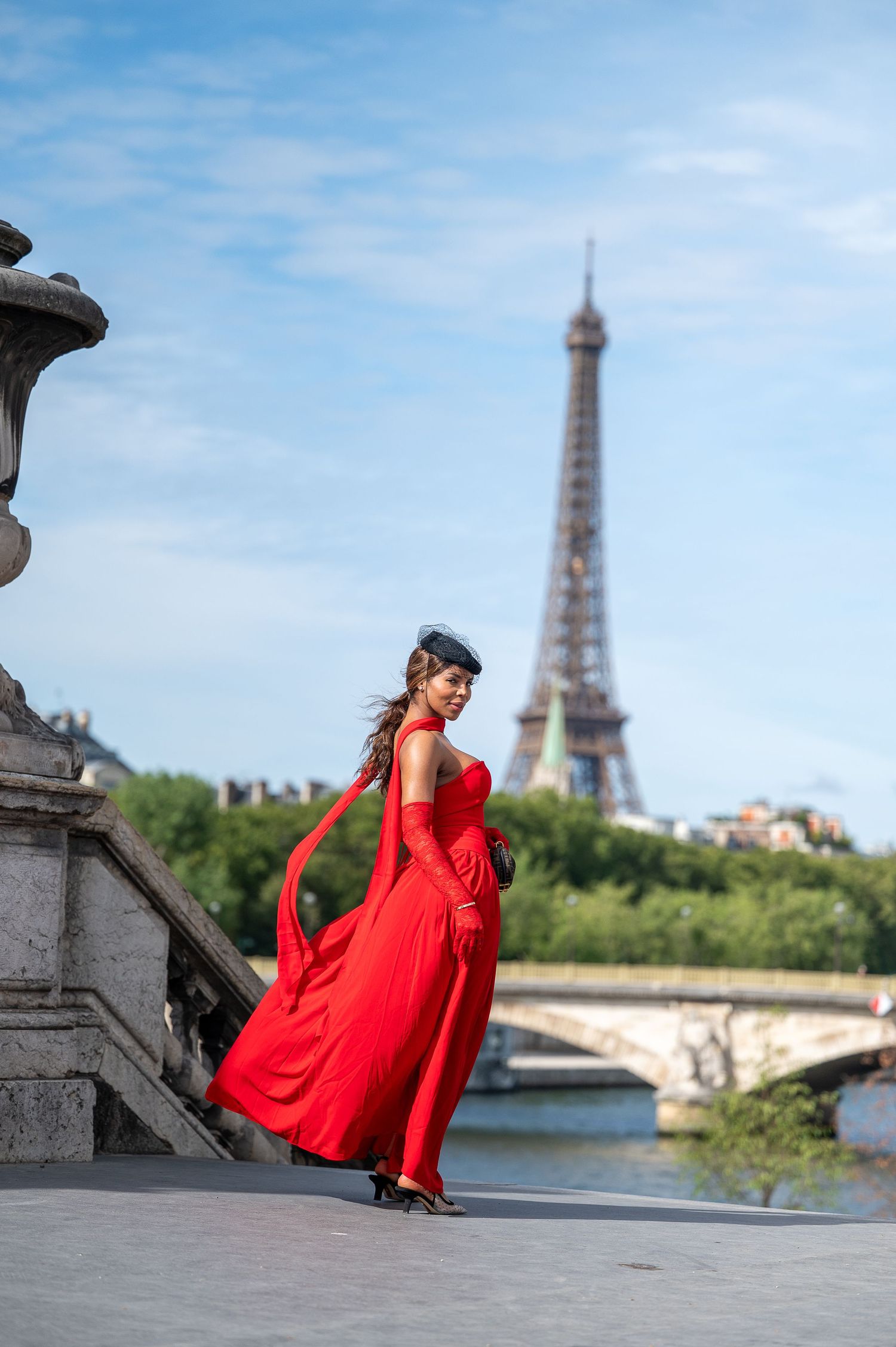 Woman in red gown and beret poses near Eiffel Tower in Paris, France on a sunny day.