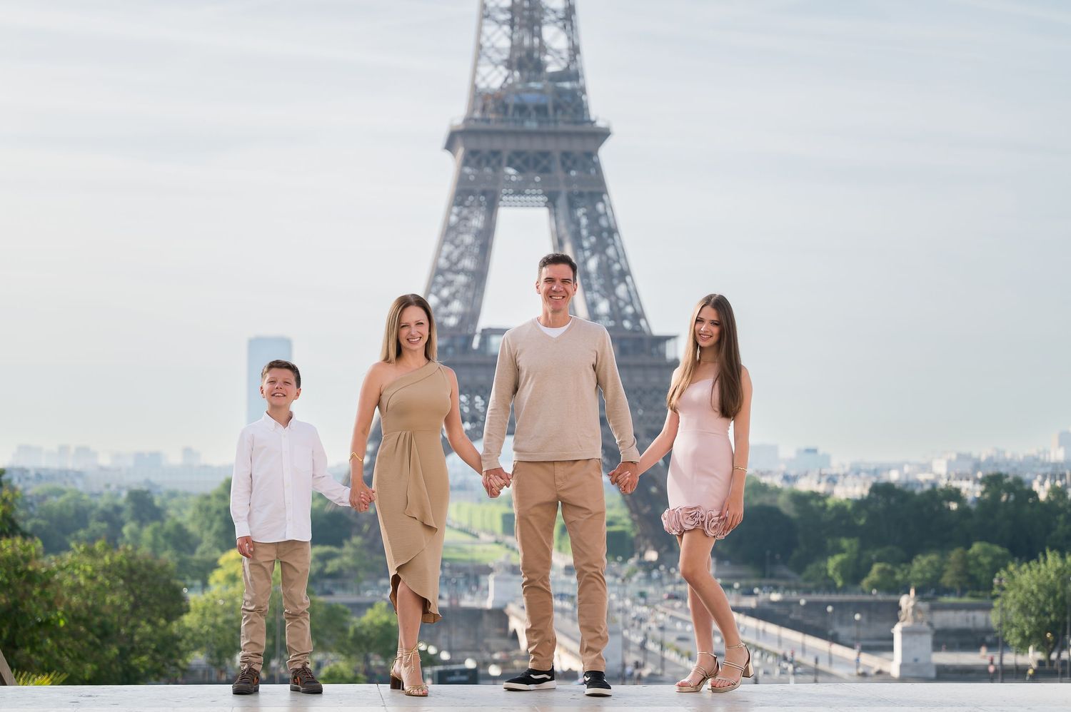 Family of four posing hand-in-hand in front of the Eiffel Tower in Paris on a sunny day.