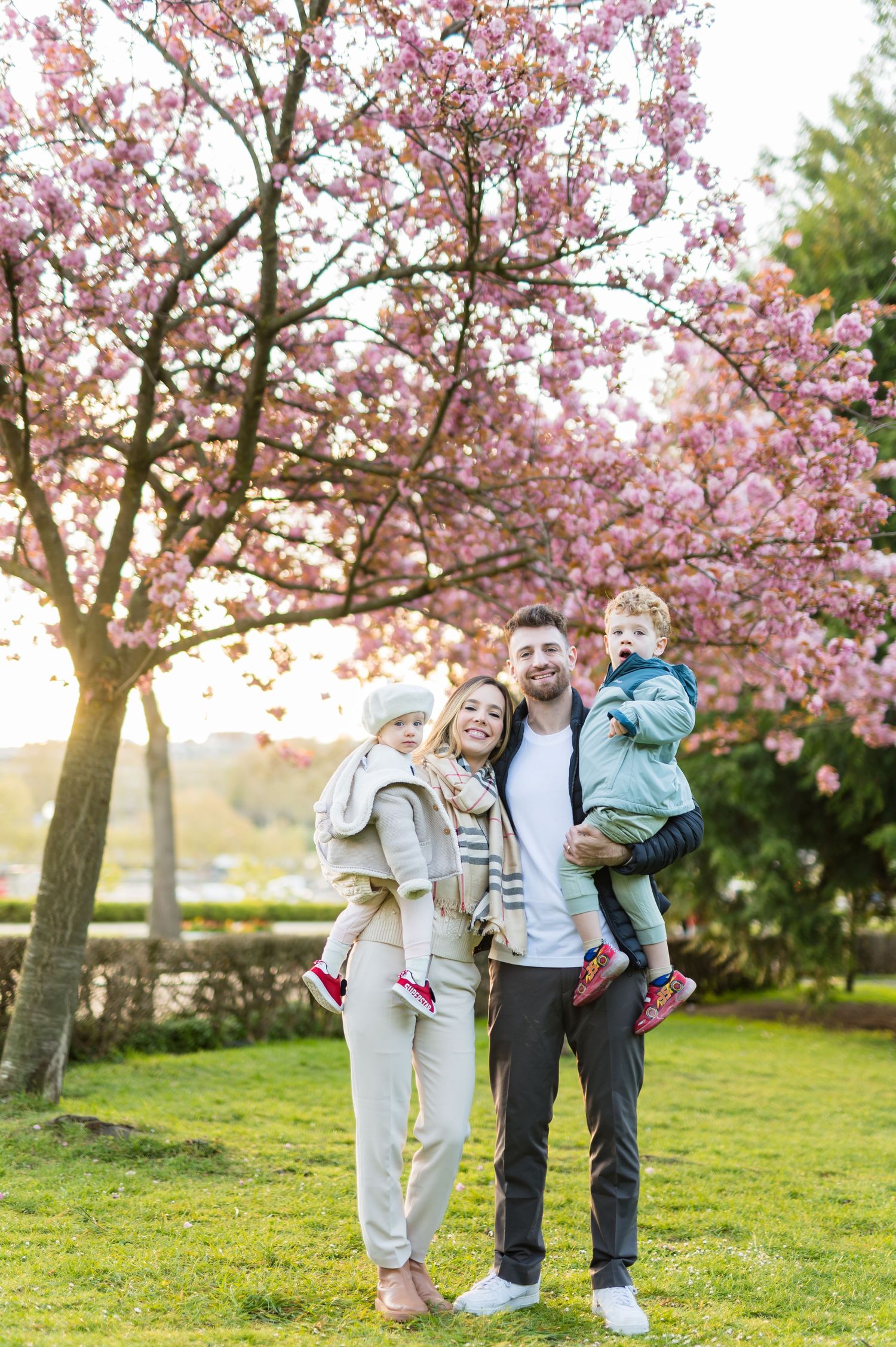Happy family of four posing under pink cherry blossom trees in a park during spring sunset.