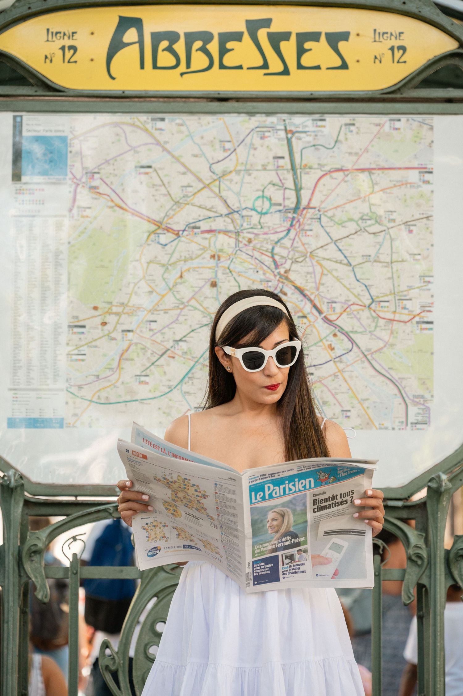 A figure in a white dress and sunglasses sits in transit while reading various maps and newspapers in Paris.