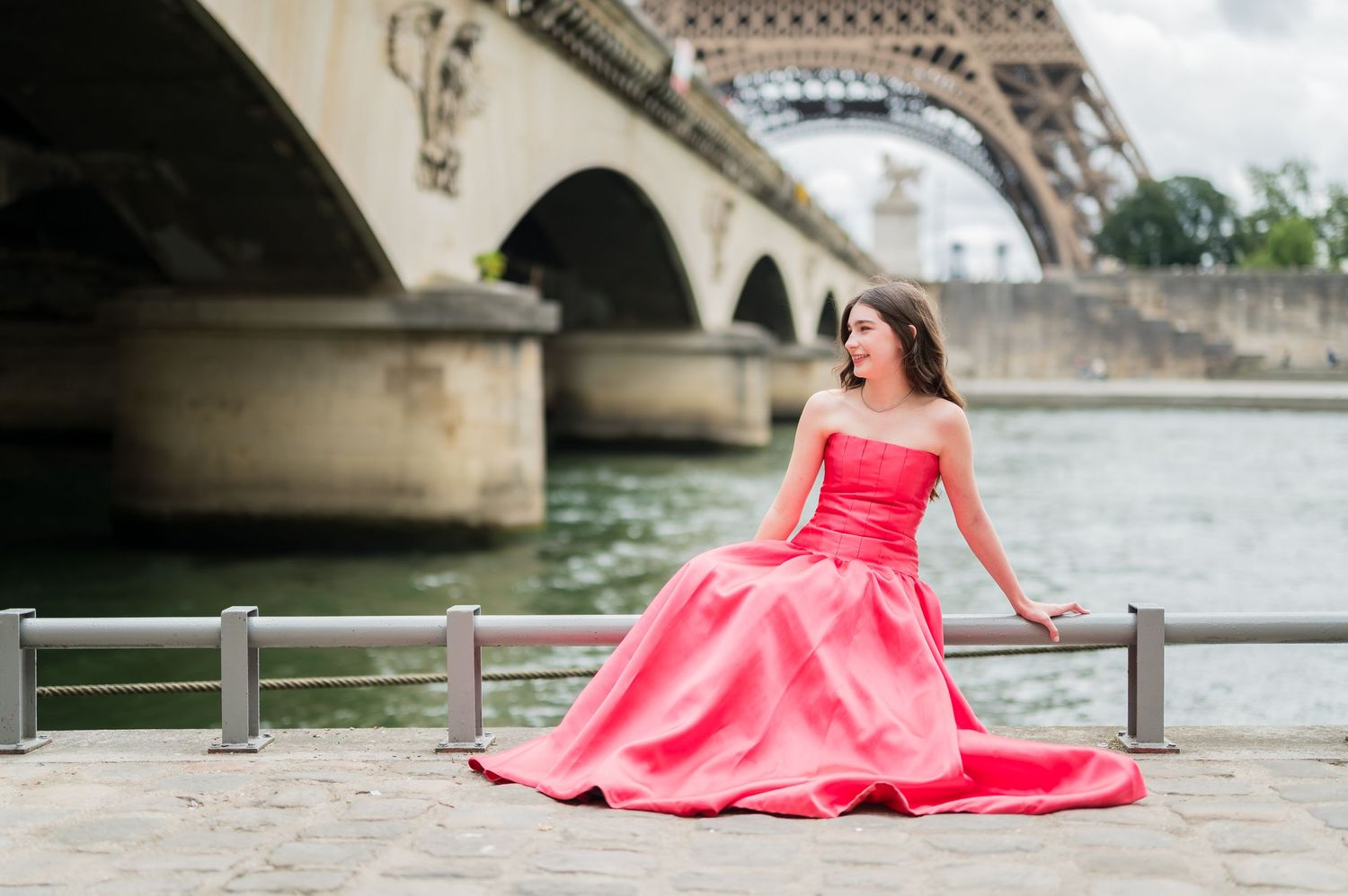 Woman in flowing coral gown poses elegantly by Seine River with Eiffel Tower in background during Paris photoshoot.