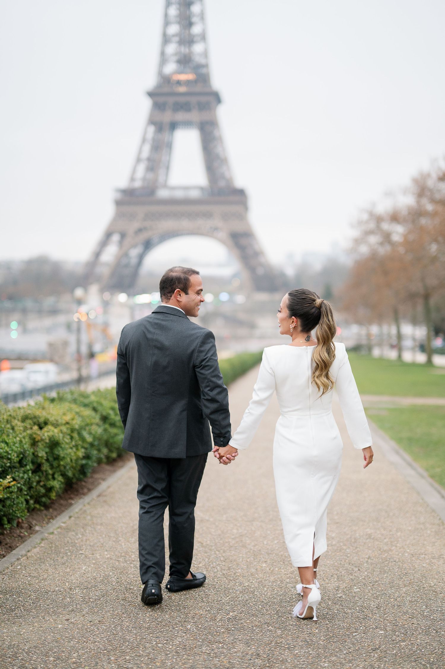A couple in wedding attire walks hand in hand along a path with the iconic Eiffel Tower in Paris visible in the distance.