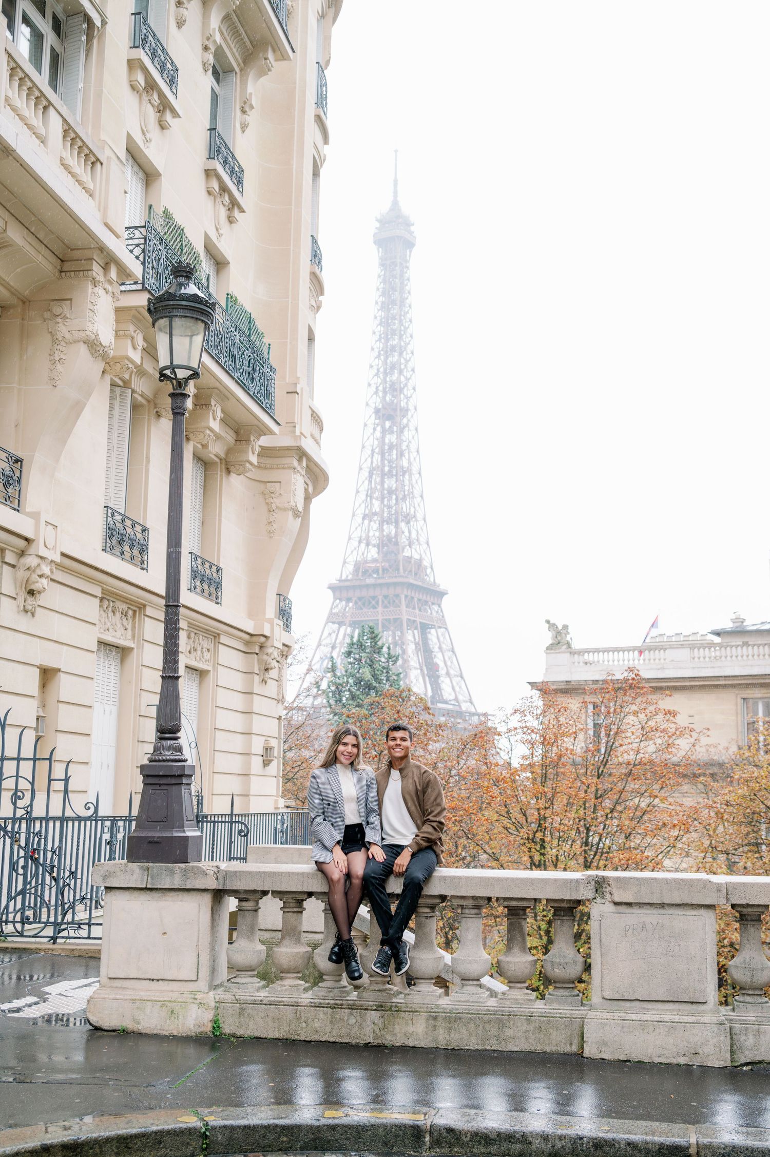 A couple sits on a stone wall with the Eiffel Tower visible between classic Parisian buildings.