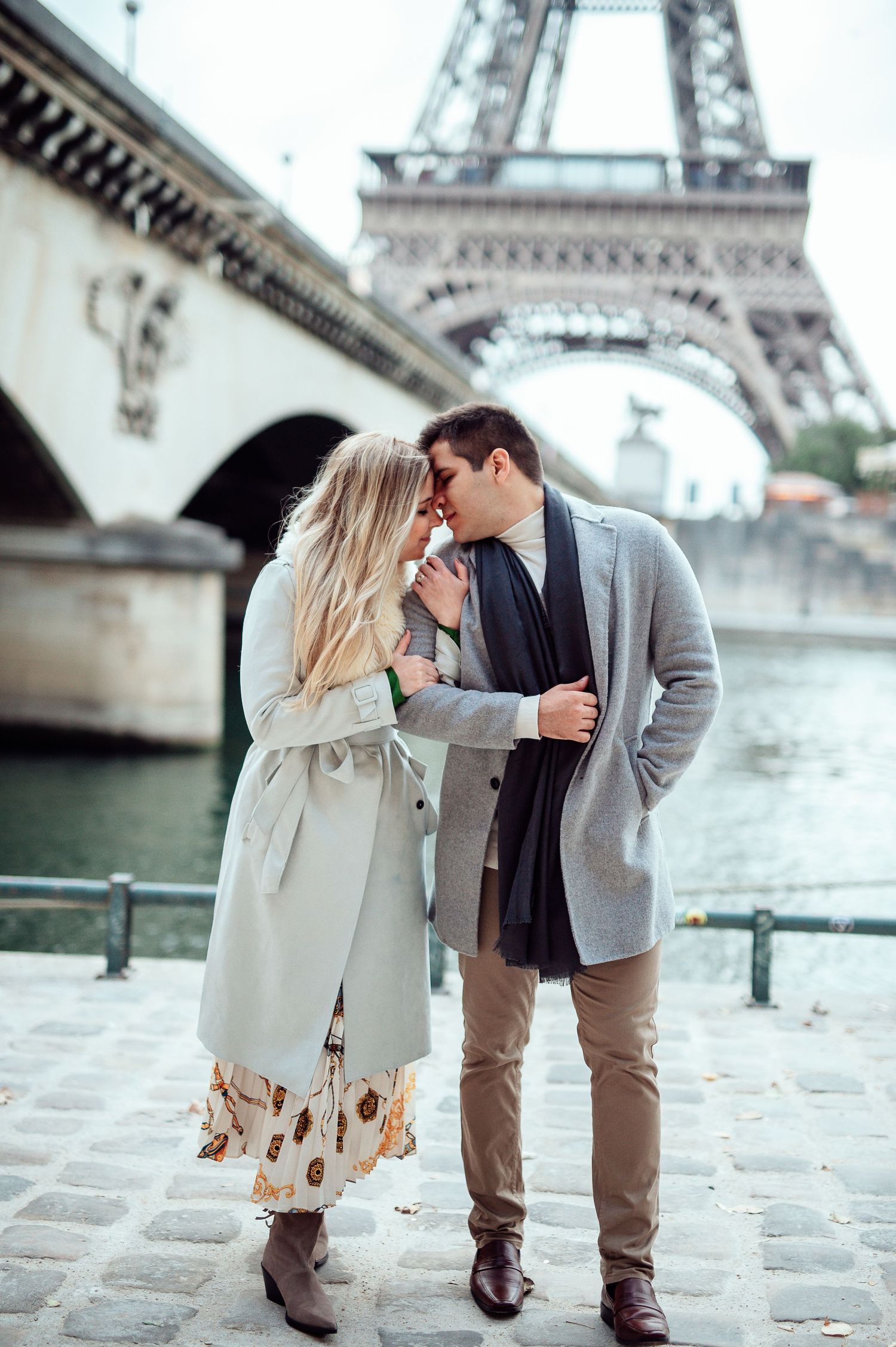 Romantic couple kissing near the Eiffel Tower in Paris, dressed in elegant fall coats by the Seine River.