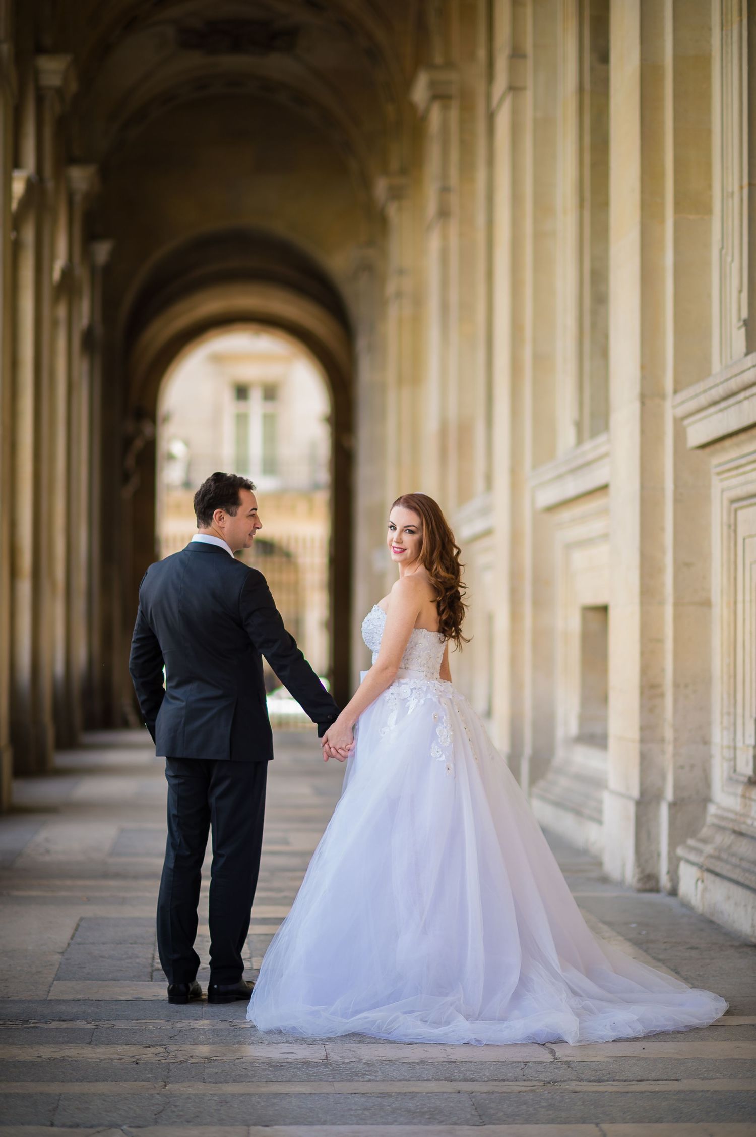 A couple shares an intimate moment in a grand arched corridor featuring classic French architecture.