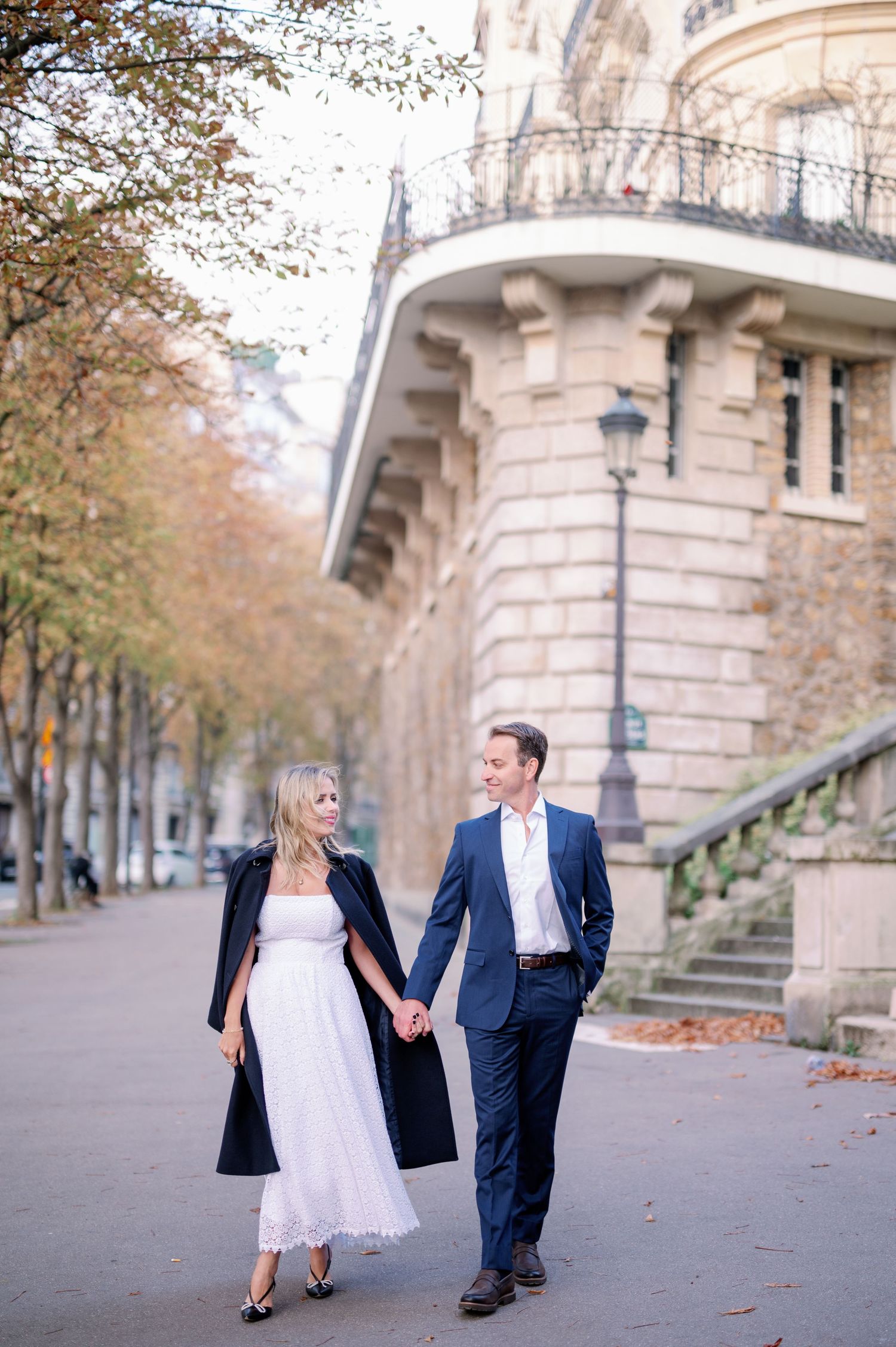 Couple holding hands walking along a Parisian boulevard in autumn, woman in white dress, man in navy suit.