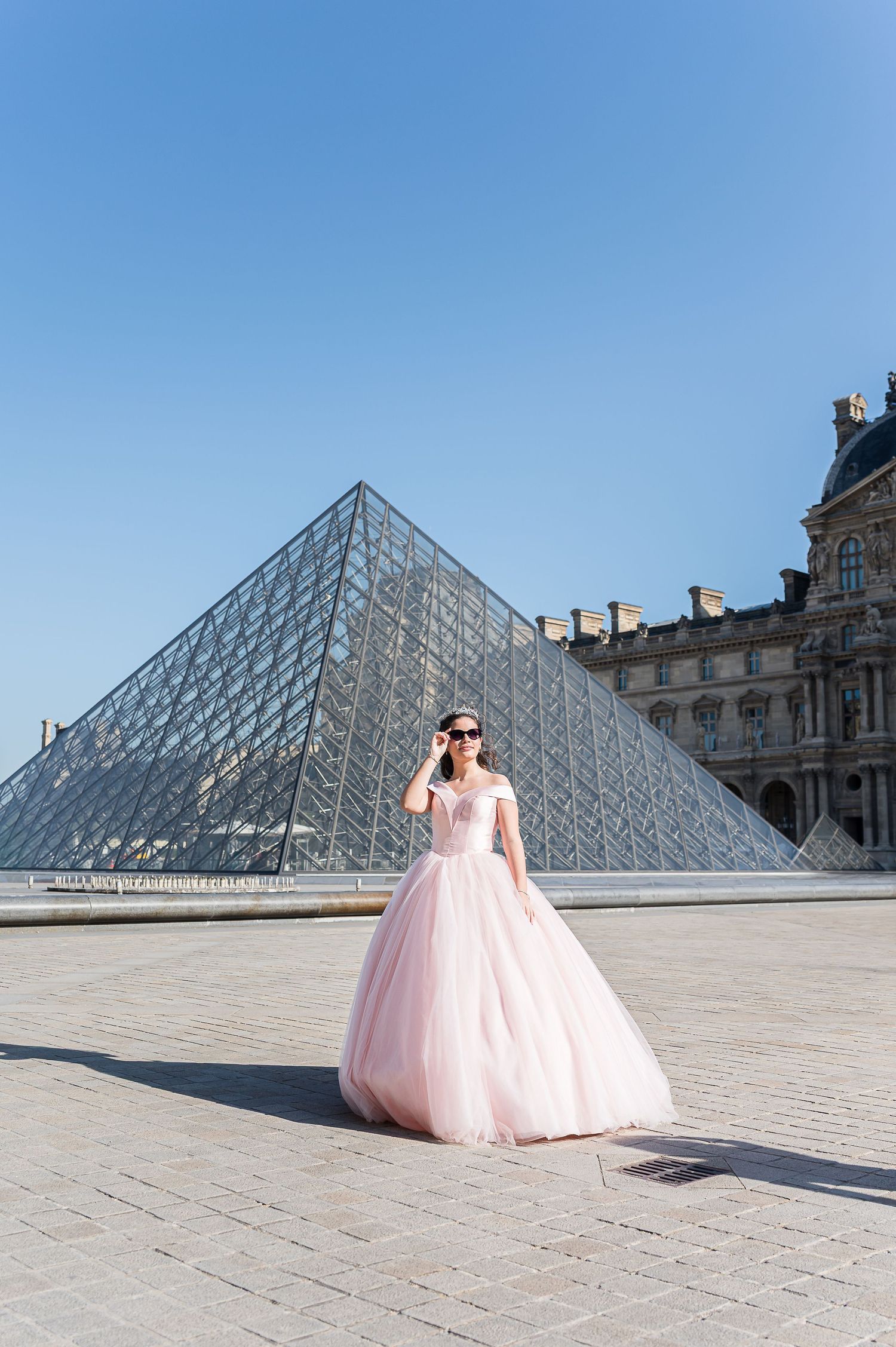 Woman in pink ballgown poses before the Louvre Pyramid in Paris on a sunny day.
