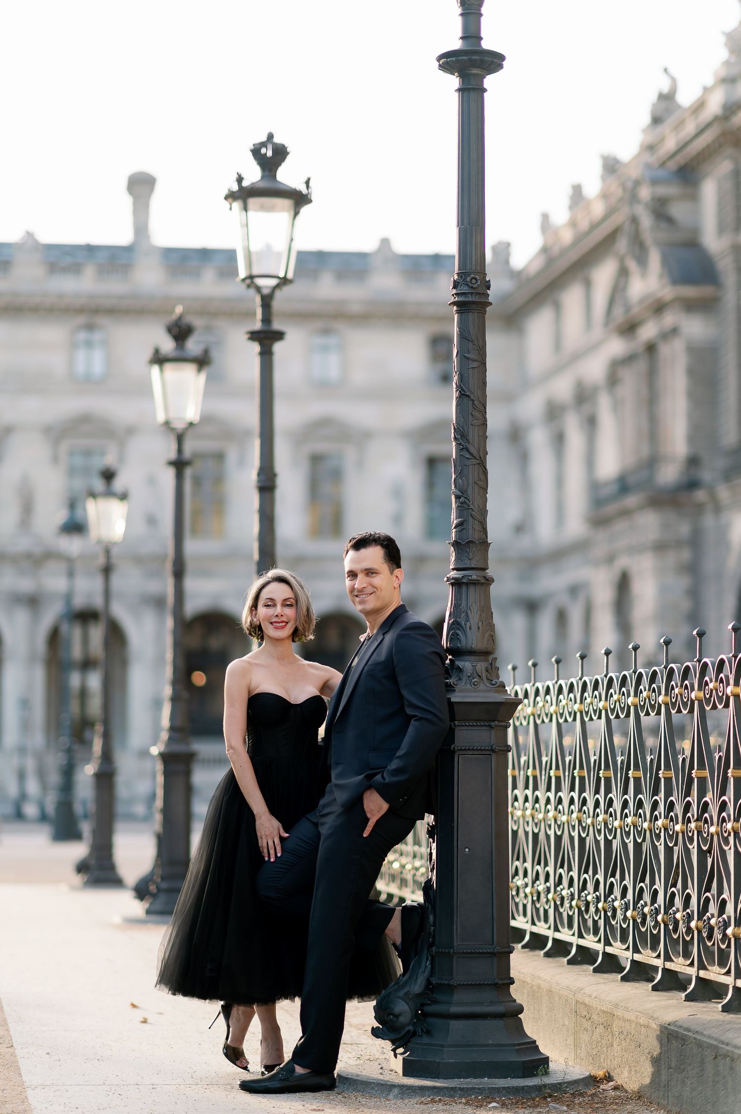 Elegant couple posing by ornate lamppost near the Louvre in Paris, woman in black tulle dress.