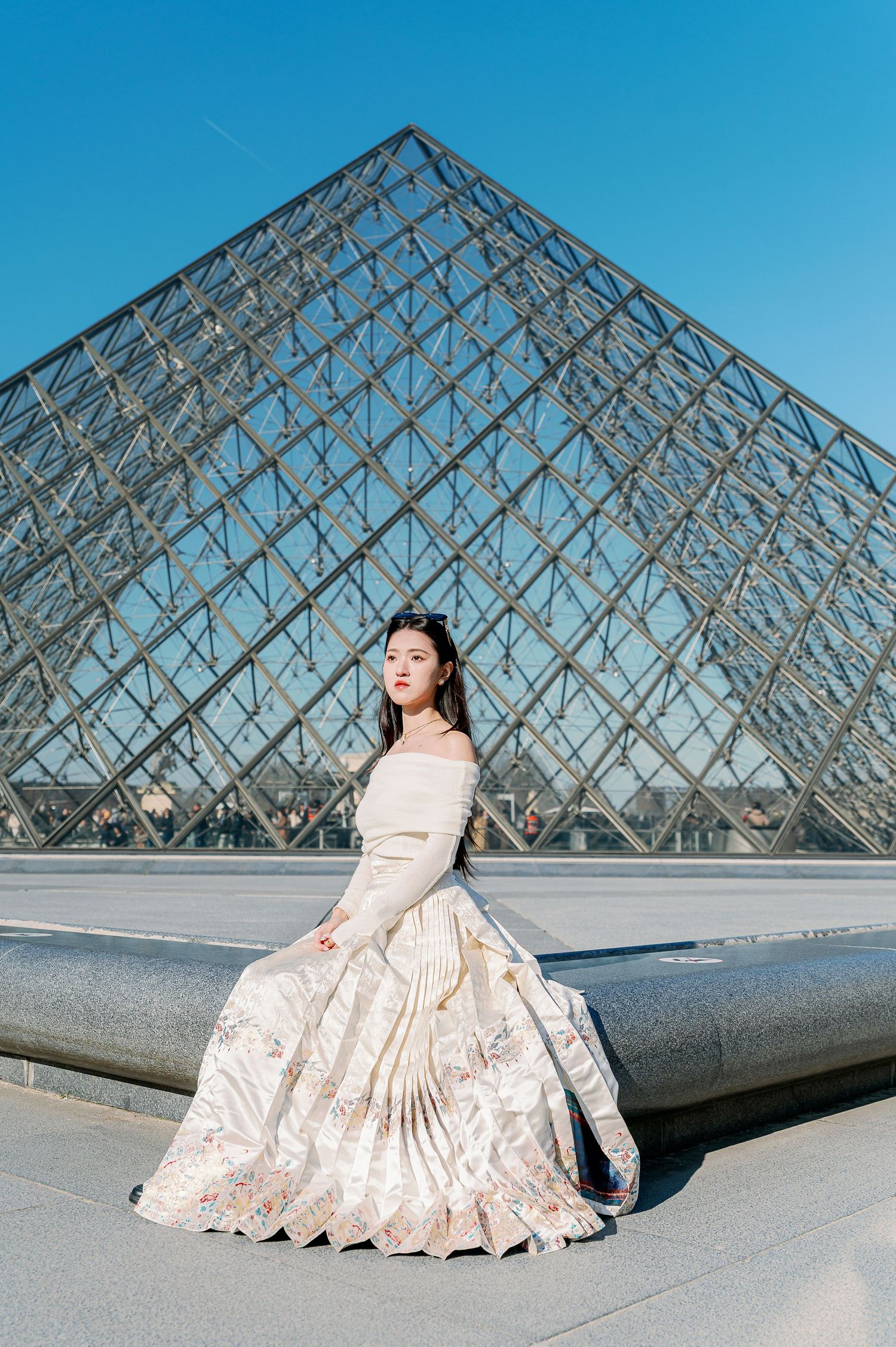 Elegant white gown photographed in front of the iconic Louvre glass pyramid in Paris.