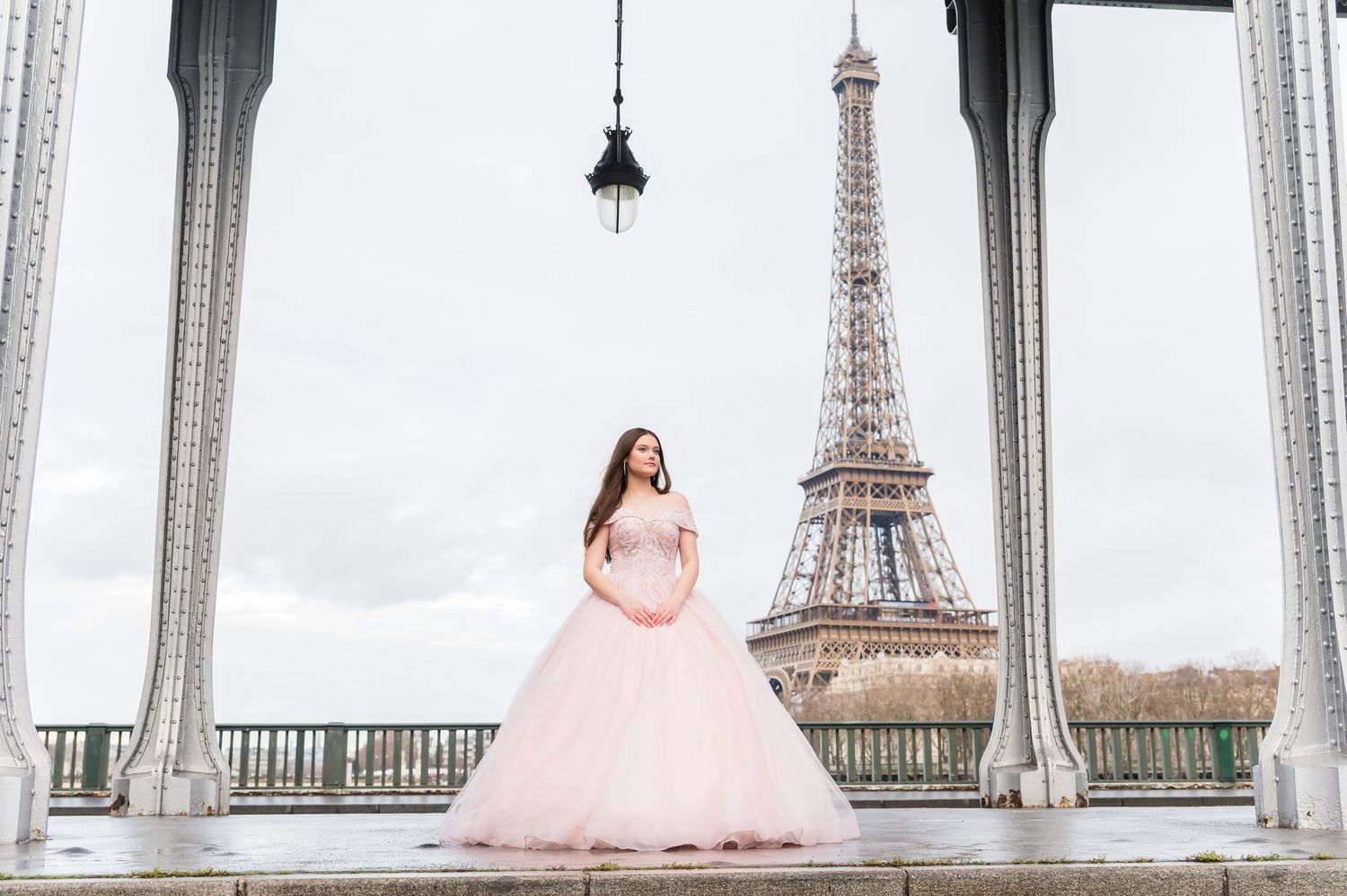 A person in a pink ballgown poses on Bir Hakeim Bridge with the Eiffel Tower visible in the background in Paris.