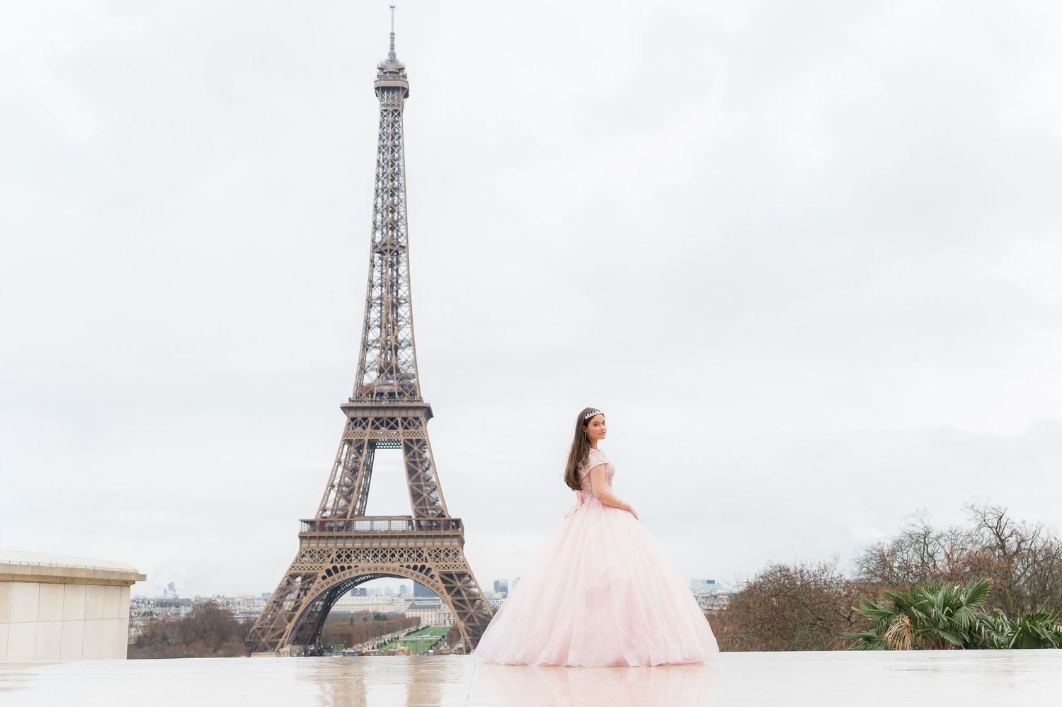 A person in a pink ballgown poses in front of the iconic Eiffel Tower in Paris on a cloudy day.