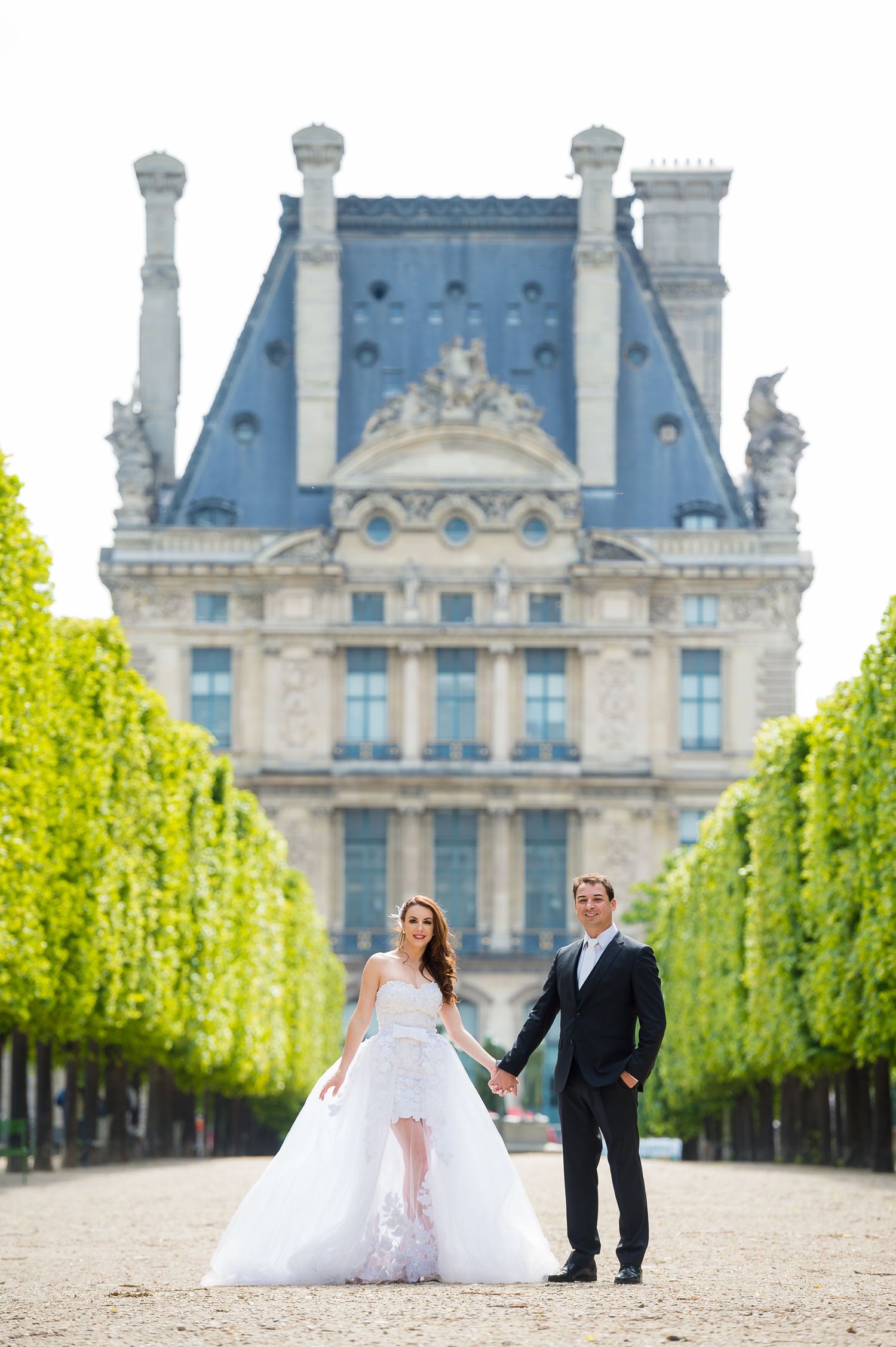 Elegant wedding couple holding hands at the gardens of the historic Louvre Museum in Paris with ornate architecture behind them.